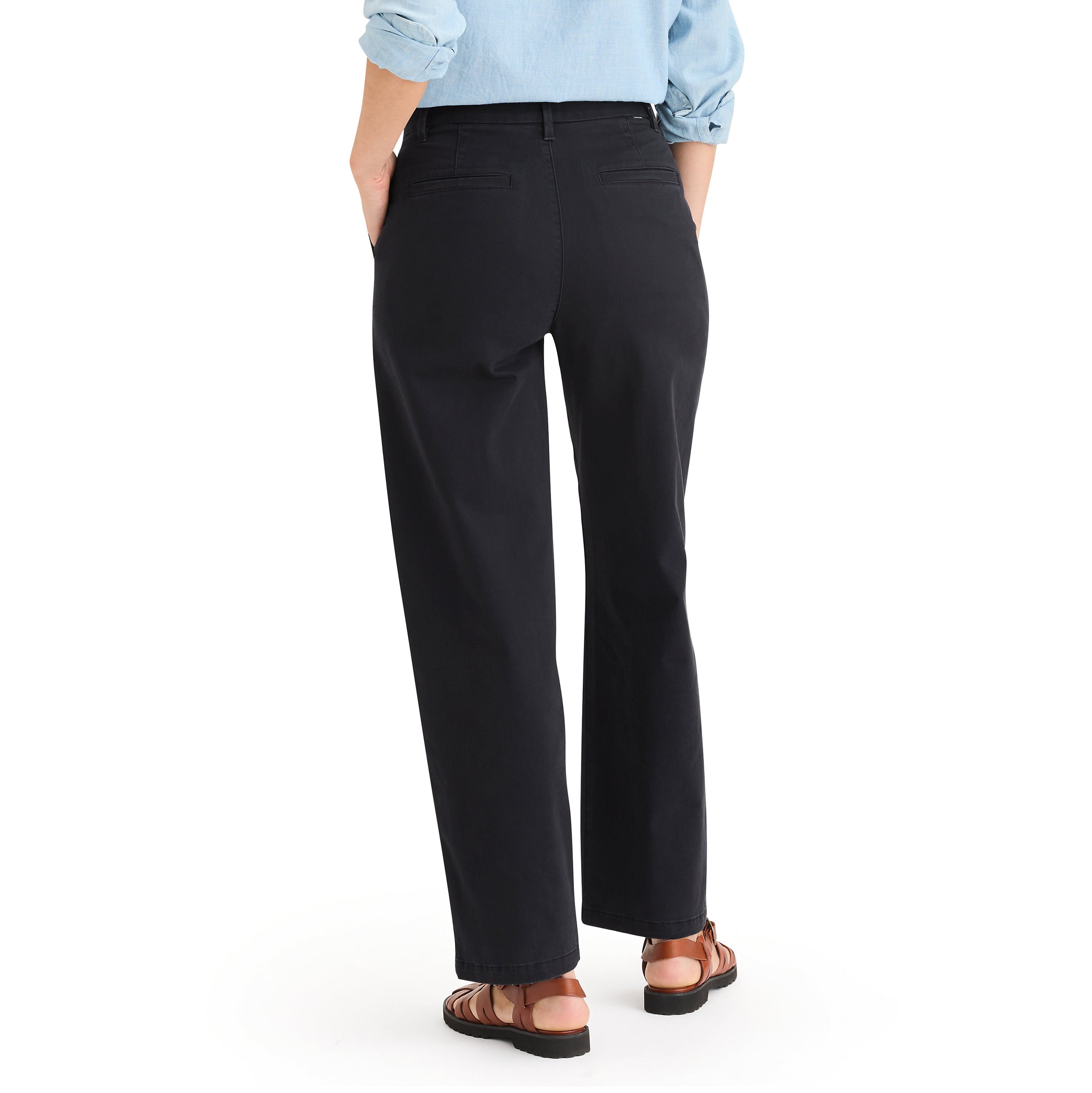 A person stands with back to camera, wearing Dockers W's Blake Chino in black recycled peached twill for a leg-lengthening look, styled with a light blue rolled-sleeve shirt and brown sandals.