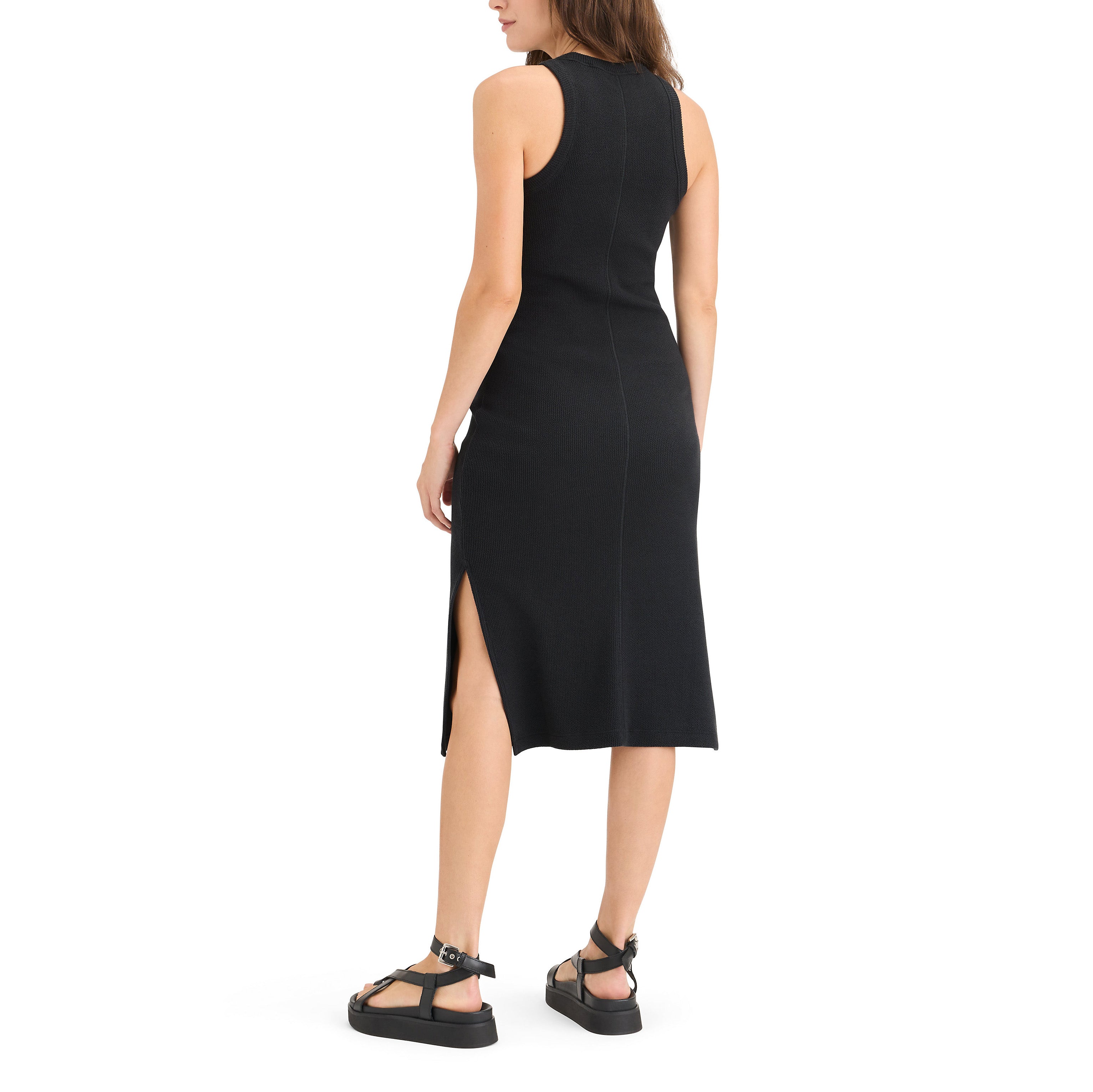 A person is wearing the Dockers Women's Loft Knit Tank Dress, a sleeveless black tank outfit with a side slit, paired with black sandals. They stand on a white backdrop facing slightly away.