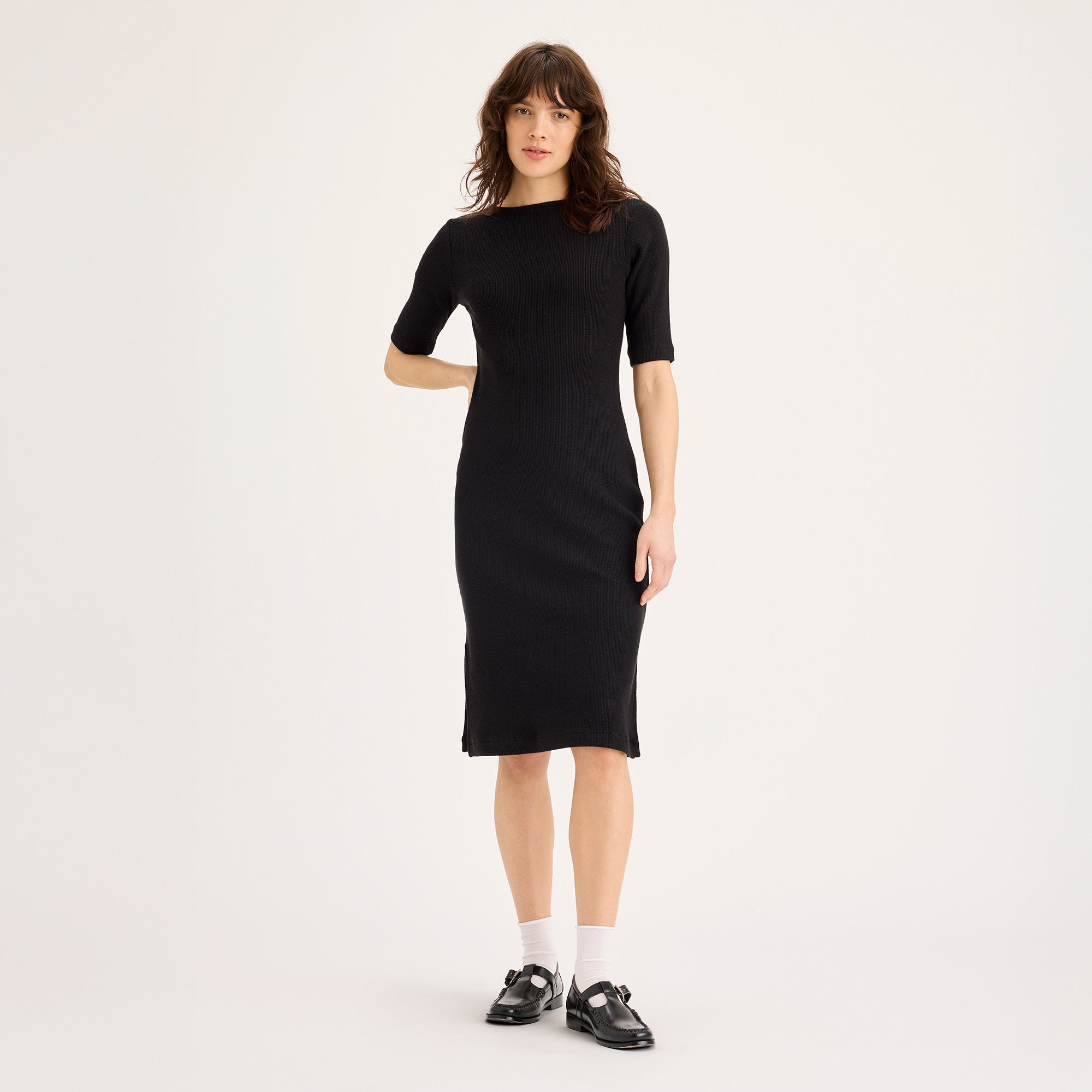 A woman stands against a plain background wearing the Dockers Winter Loft Knit Dress in Beautiful Black, paired with white socks and black shoes, her arms relaxed at her sides.