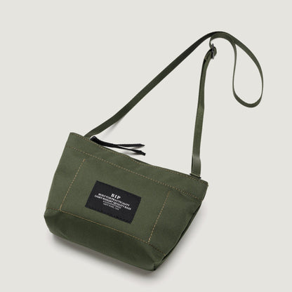 Olive green canvas Zipper Pouch Mini crossbody bag from Bags in Progress with an adjustable strap and a black label on the front.