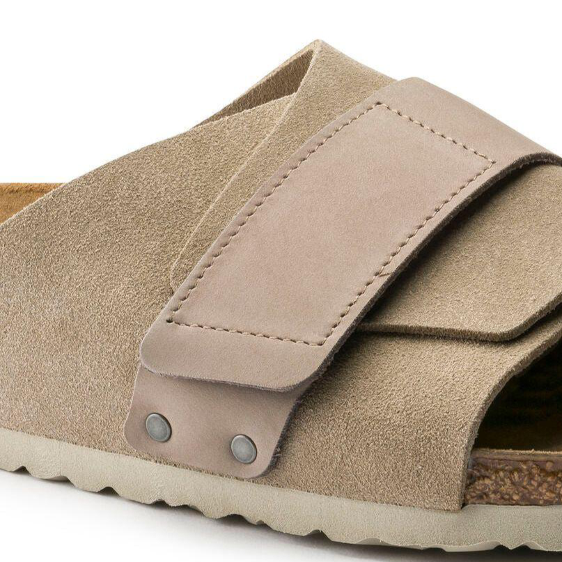 Close-up of the Birkenstock Kyoto Suede sandal in taupe, featuring a tan suede upper with a wide overlapping strap secured by metal rivets, a cork-latex footbed, and textured rubber sole.