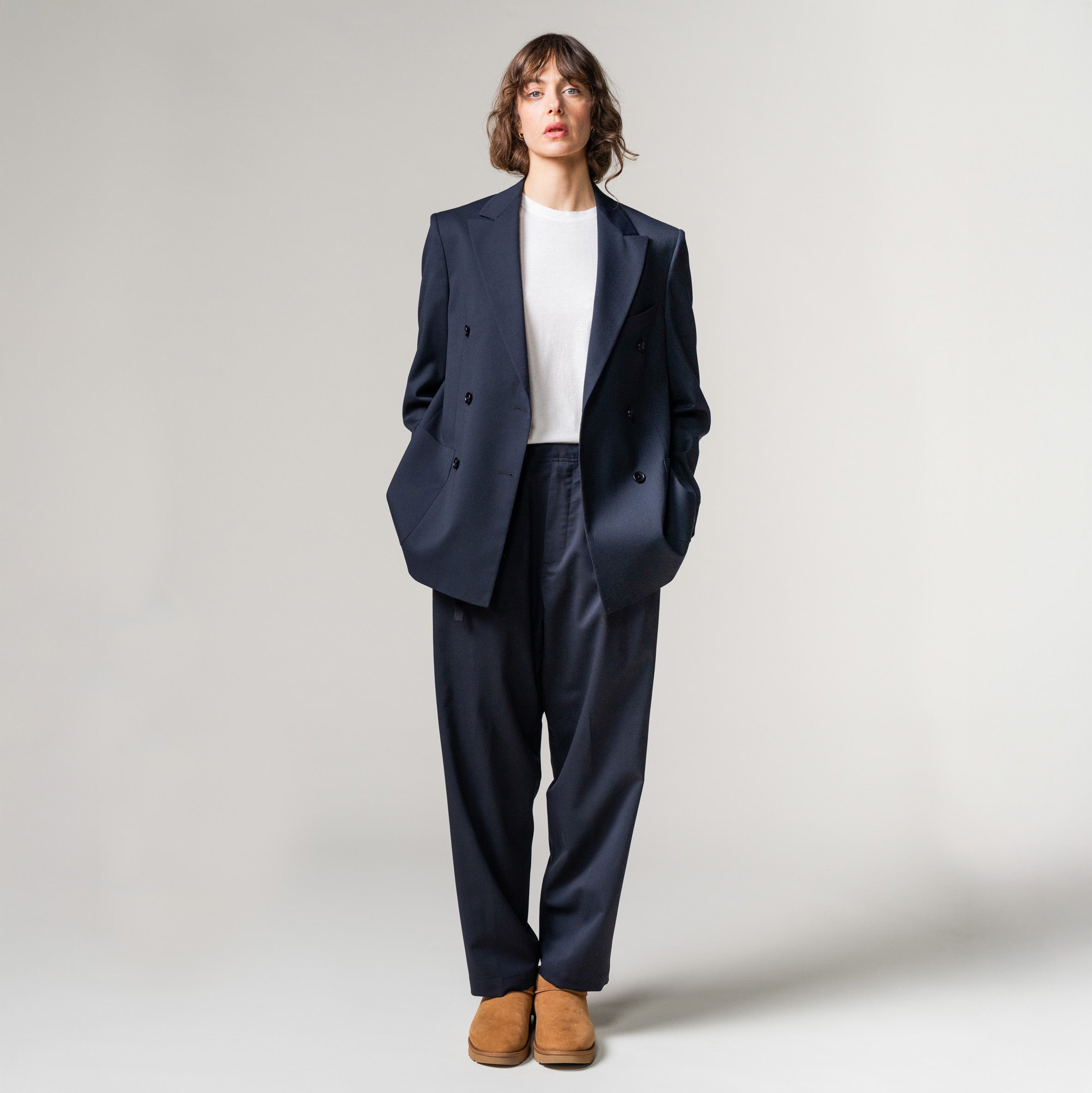A person dons an oversized navy suit with a white shirt and brown shoes, standing against a plain backdrop. The Seven Gauge Double Breast Blazer in Tropical Wool enhances the ensemble's sophistication.