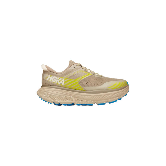 The Hoka M Stinson ATR 6 in DOTN is shown in side profile on a white background, featuring a thick sole, beige upper with yellow accents, and a blue outsole—delivering aggressive performance and versatile comfort for every run.