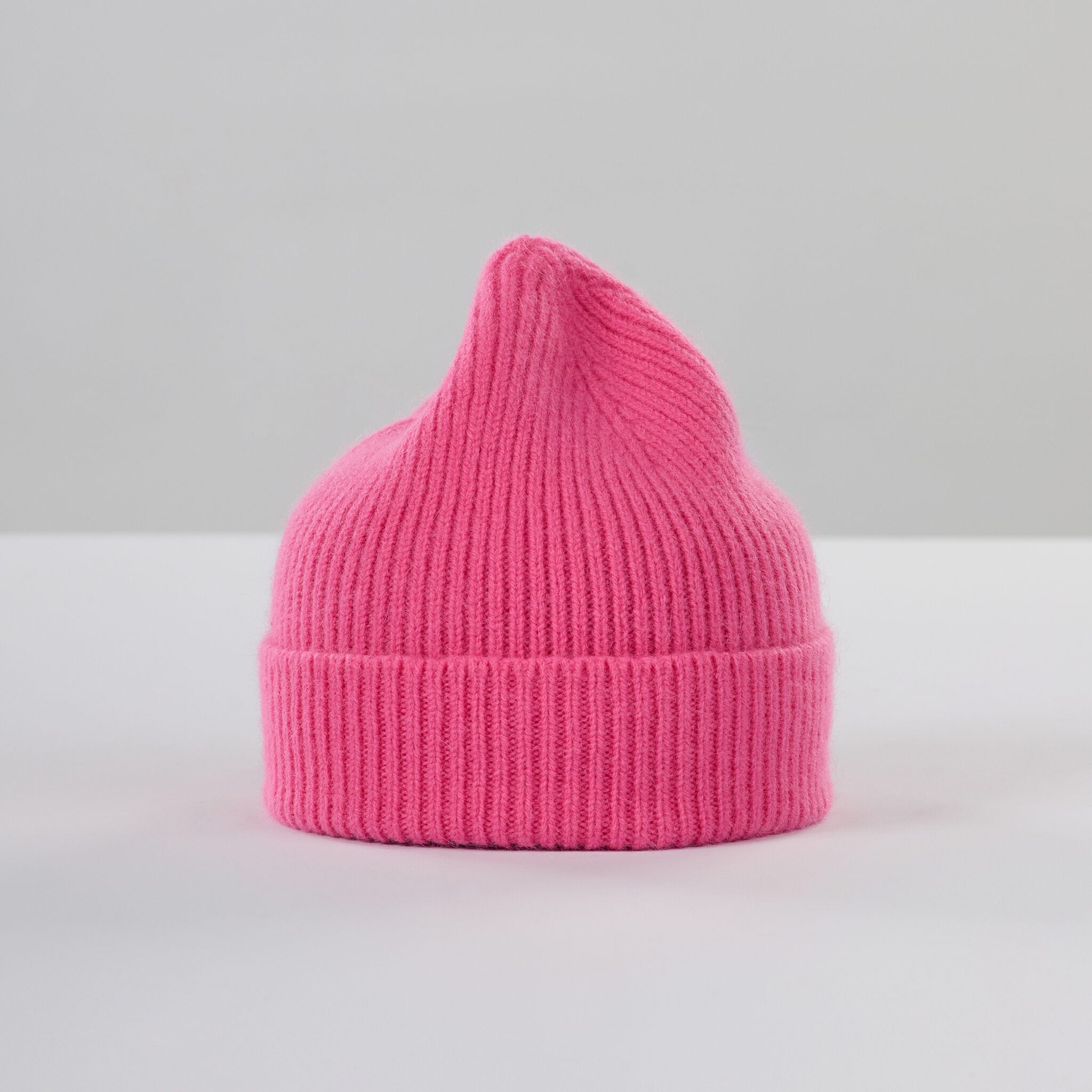 The Le Bonnet Beanie in Bubblegum, featuring a ribbed knit lambswool design and slightly pointed top, stands upright on a white surface against a plain light gray background.