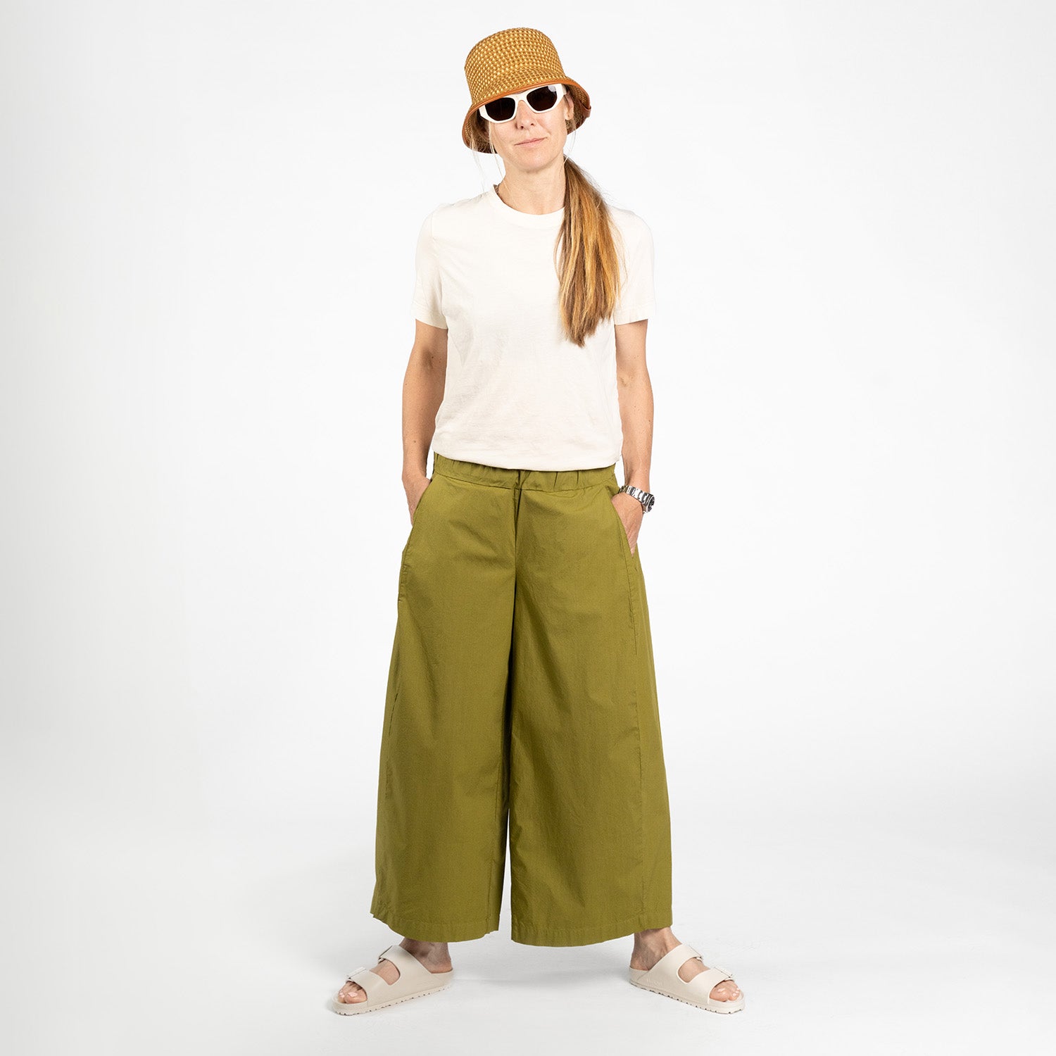 A person wears a beige t-shirt, Labo Art Panta Storto Pant in Clara, sandals, a brown textured bucket hat, and white sunglasses, standing hands in pockets against a plain white background.