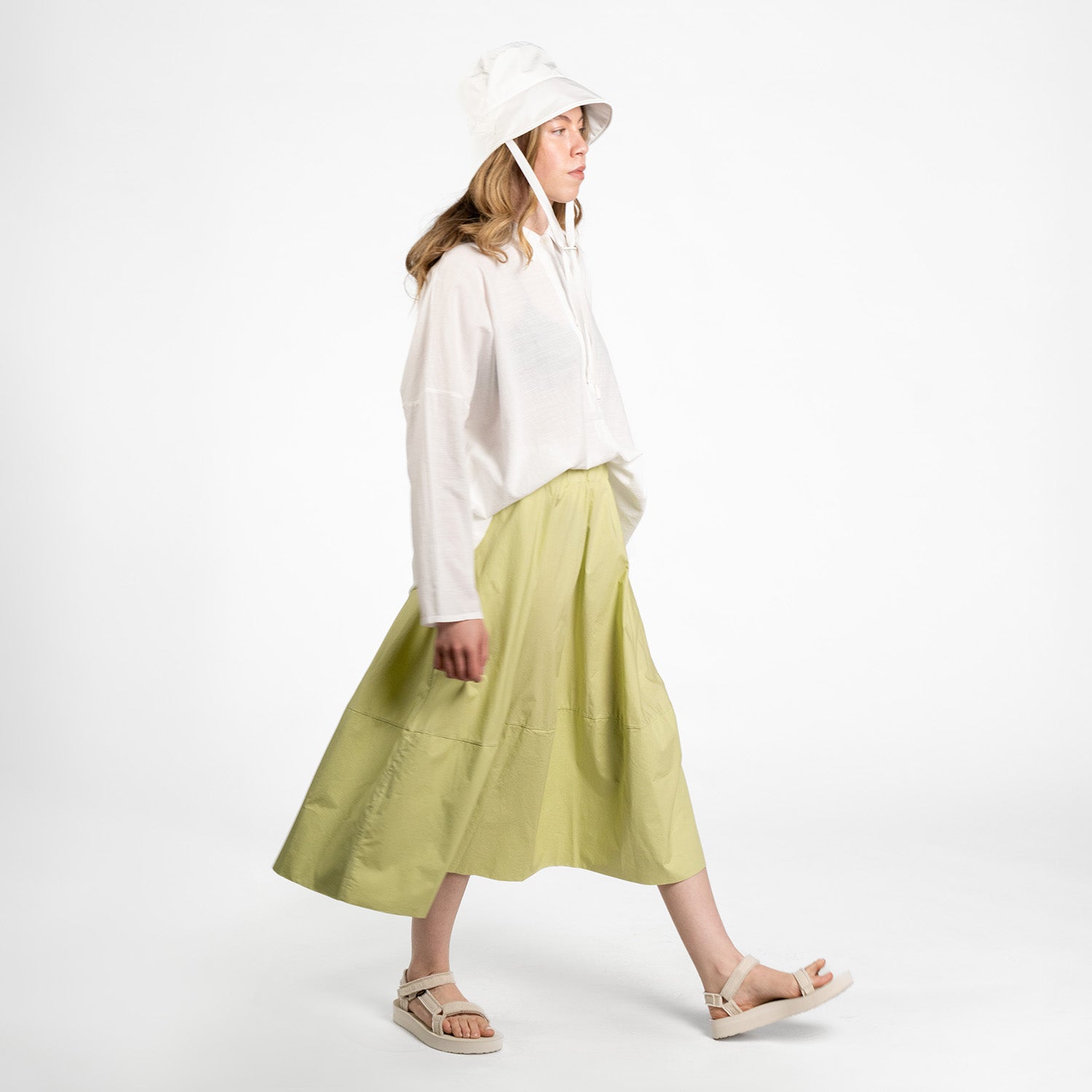 A person in a white hat, white long-sleeve top, Labo Art's Gonna Gea Skirt in light green by Clara, and beige sandals walks against a plain white background.