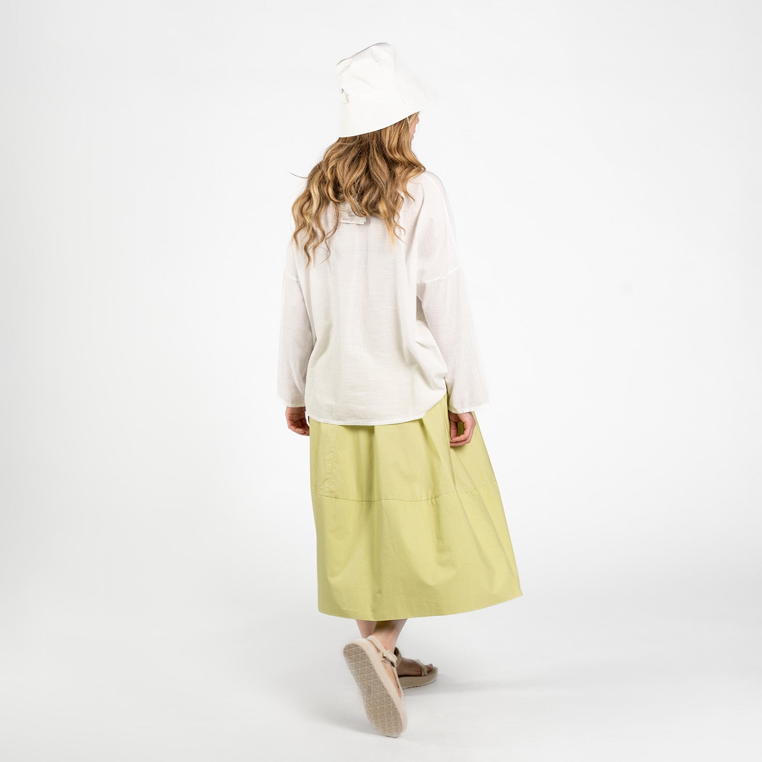 A person wearing a white long-sleeve shirt, Labo Art Gonna Gea Skirt in Clara, white bucket hat, and sandals stands facing away against a plain white background.