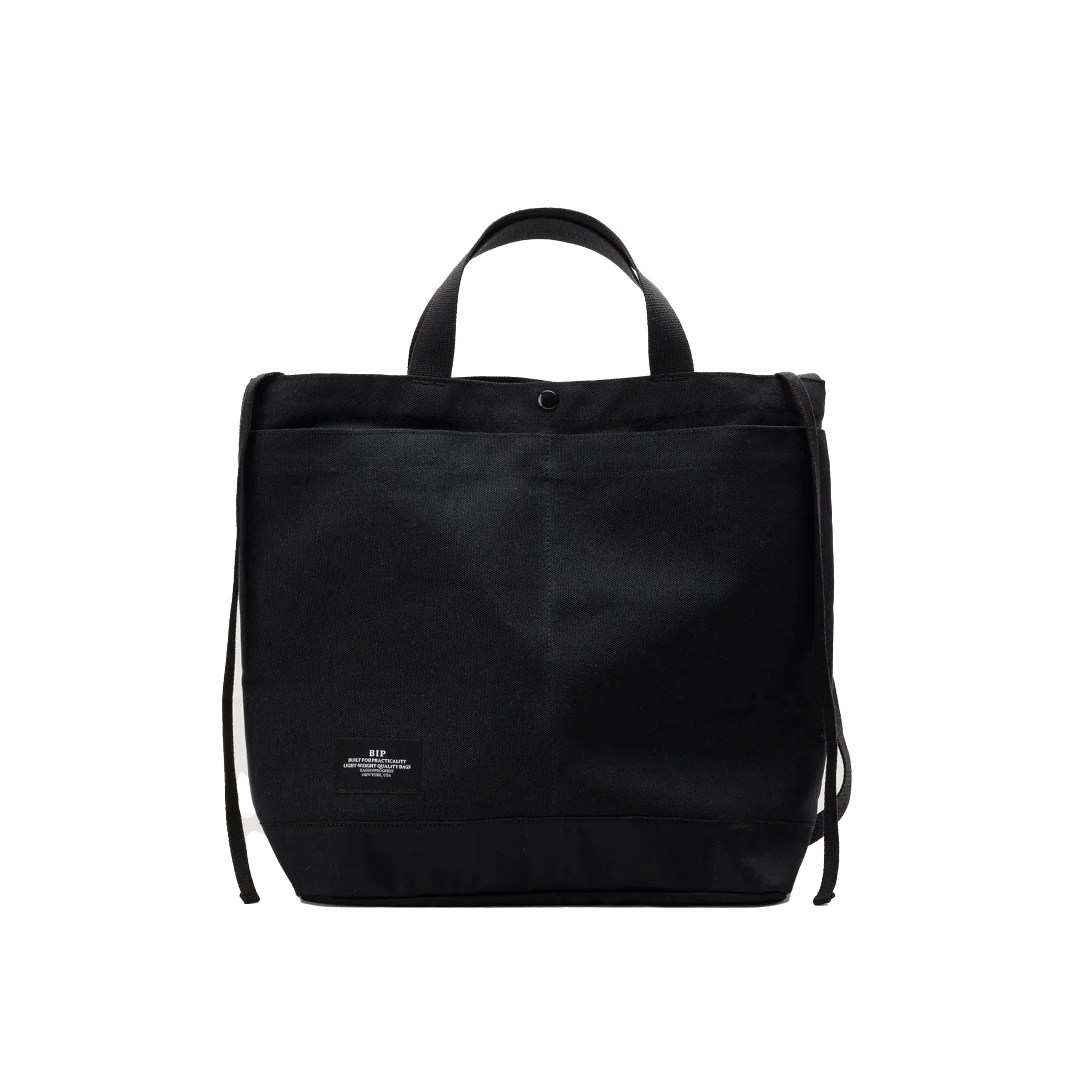The Bags in Progress New York Tote Medium is a black rectangular cotton canvas bag made in Japan, featuring short handles, an adjustable shoulder strap, and a small white label at the bottom front center.