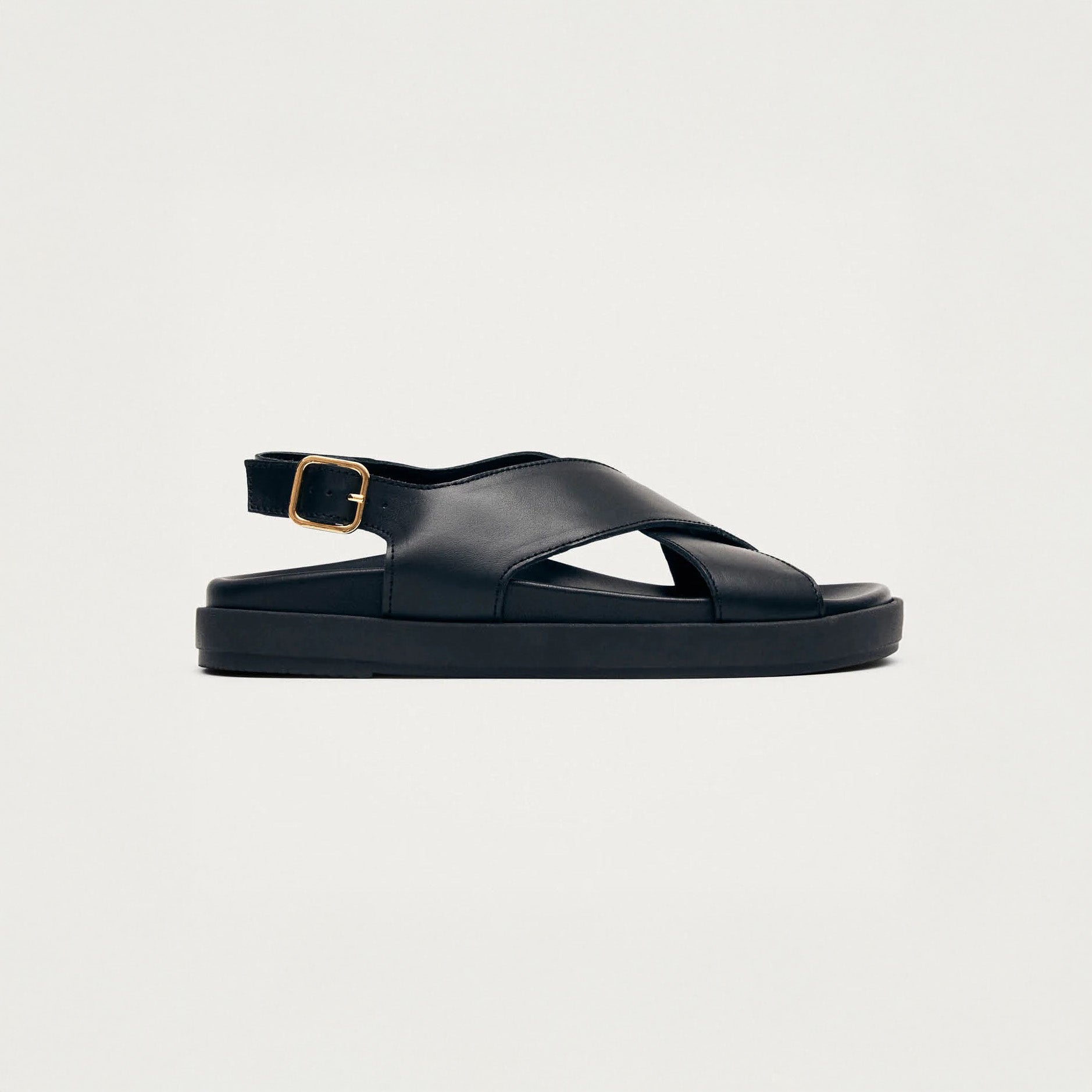The Alohas Nico Sandal in black showcases thick soles, crisscross leather straps, and a gold ankle buckle, styled as a comfy crossover slingback on a simple light backdrop.
