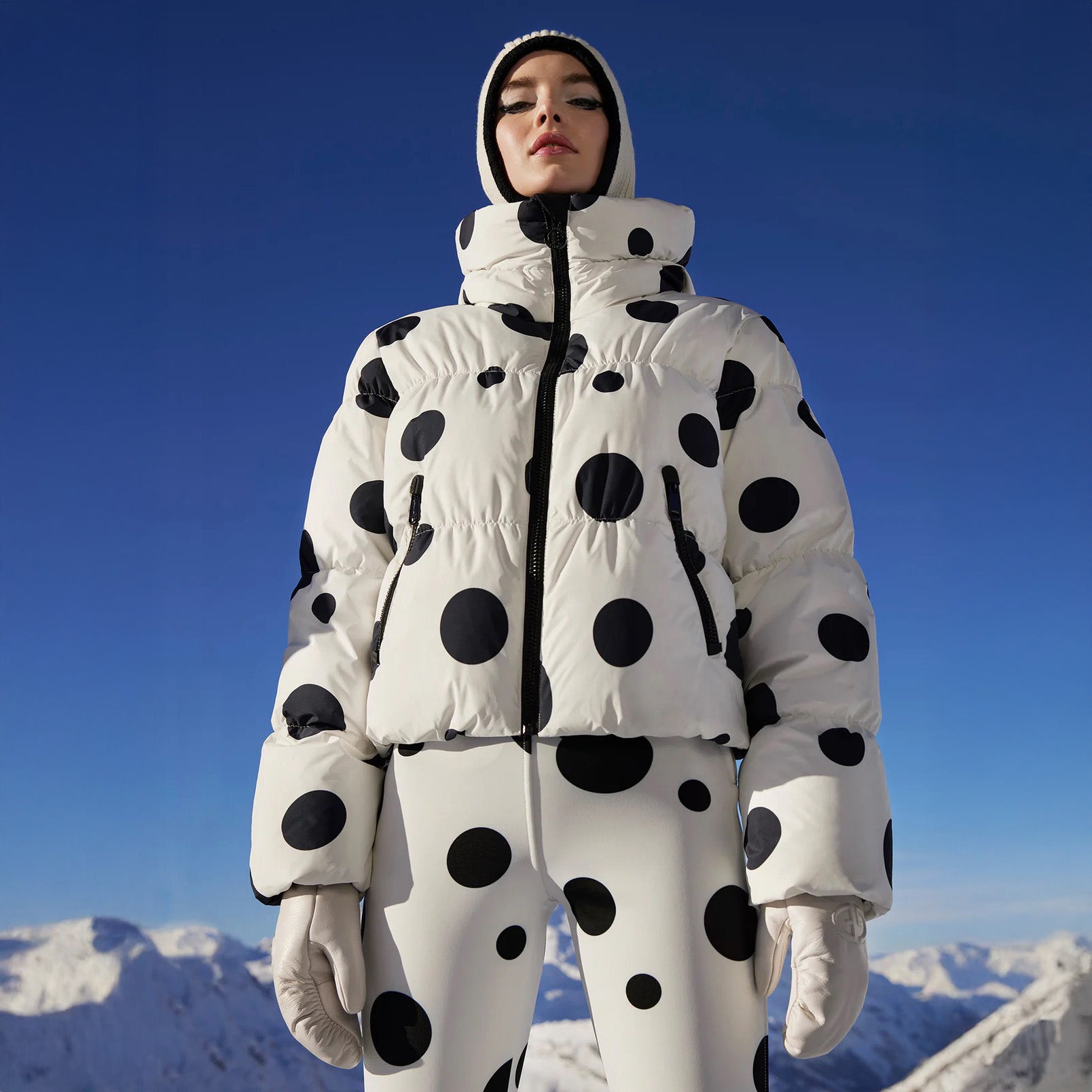 A person wearing the Goldbergh Grand Ski Jacket in Dot Cream stands in a snowy mountain landscape under a clear blue sky.