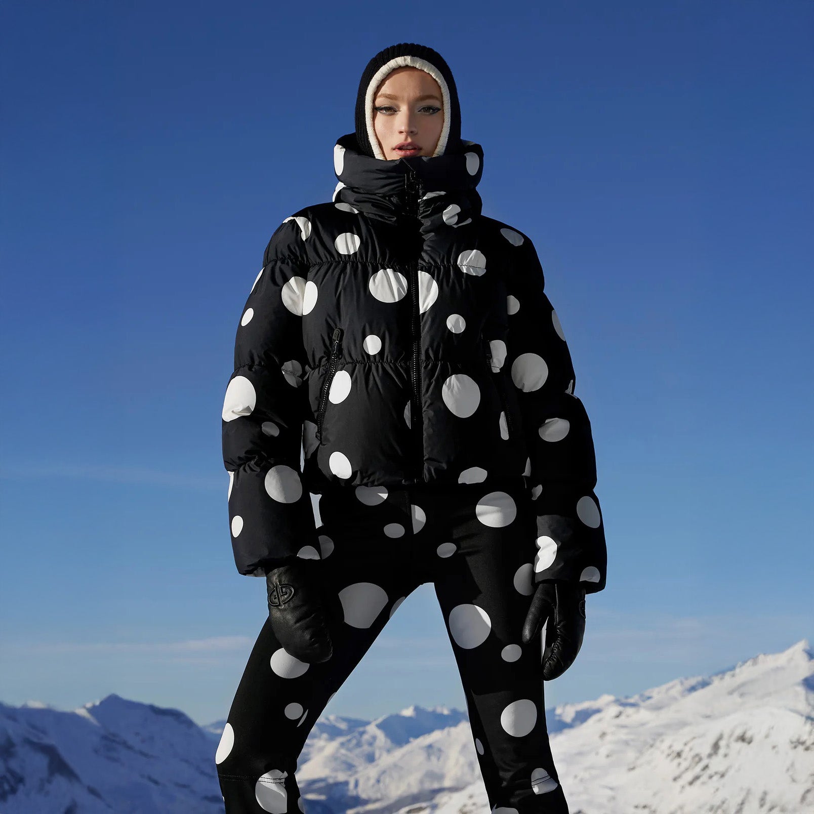 A person confidently stands in a snowy mountain landscape, wearing Goldbergh's Grand Ski Jacket, Dot Black, which features waterproof fabric providing insulation against the crisp winter chill.