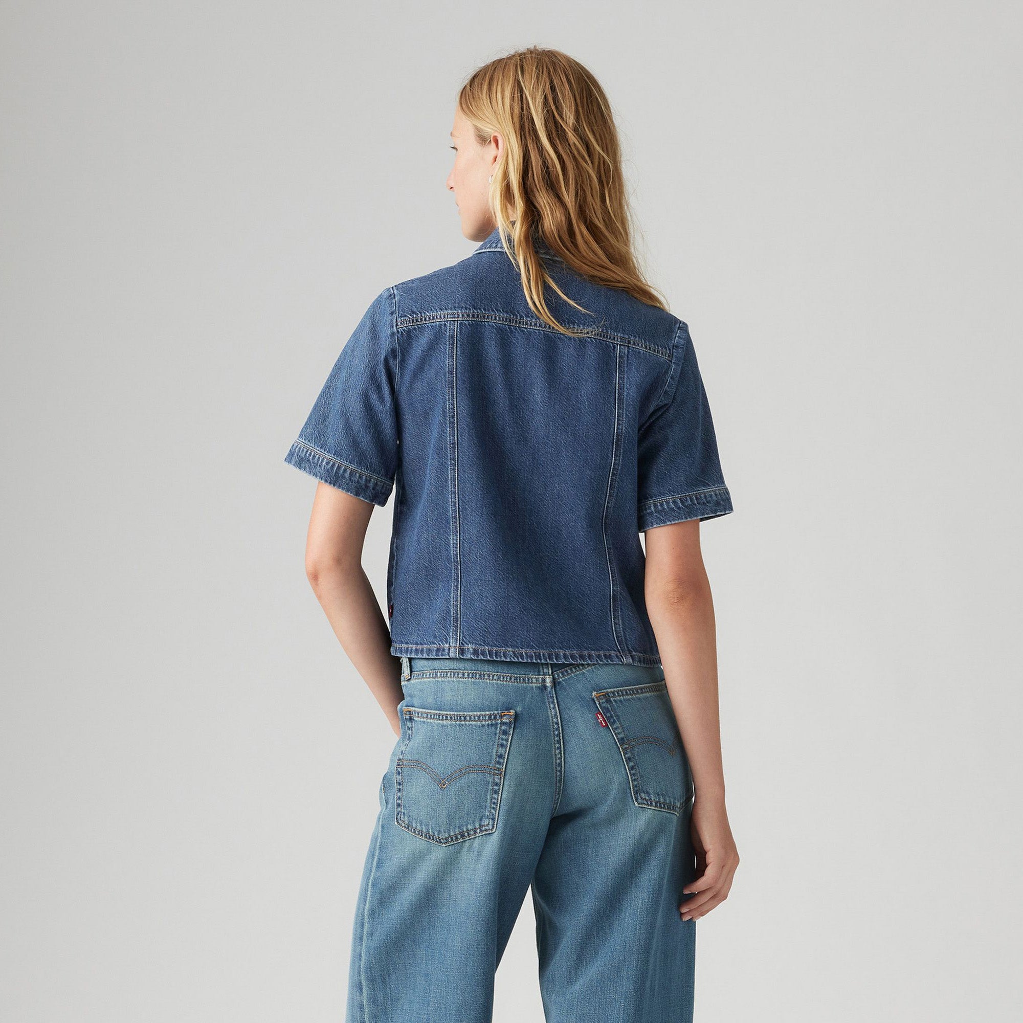 Woman with long blonde hair seen from behind, wearing the Levis Rinoa Camp Shirt and standard blue jeans against a plain background.