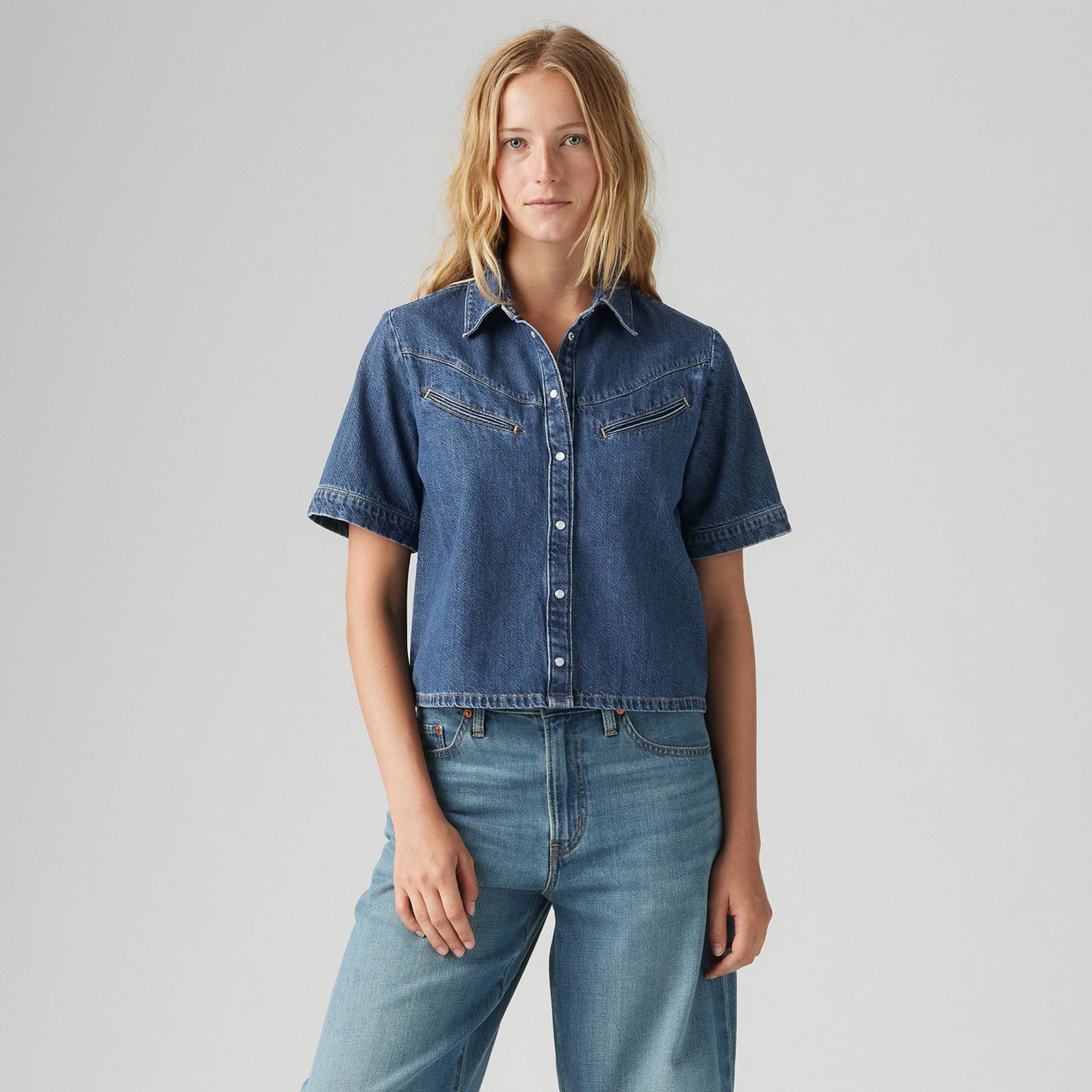 A woman faces forward in a Levi's Rinoa Camp Shirt—short-sleeved, button-up denim—paired with light blue jeans, capturing an Americana classic style against a plain light background.