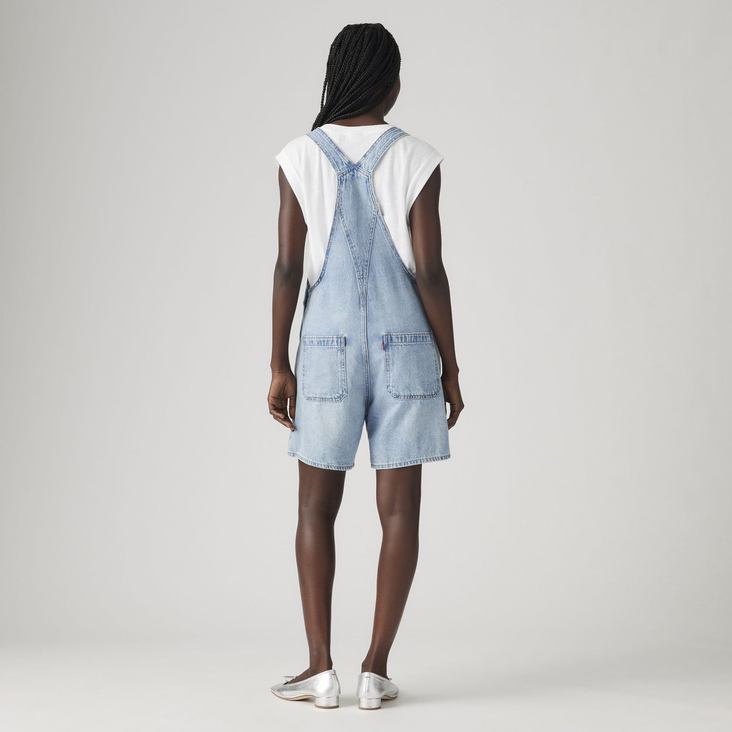 A person faces away, wearing a relaxed fit white short-sleeve shirt, Levi's Utility Shortall in light blue denim with a mid-length cut, and white shoes, set against a plain light background.