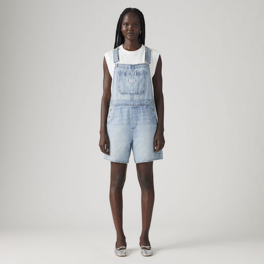 A person stands facing forward, wearing a white t-shirt and Levis Utility Shortall in light blue denim with a relaxed fit, paired with silver shoes against a plain light background.