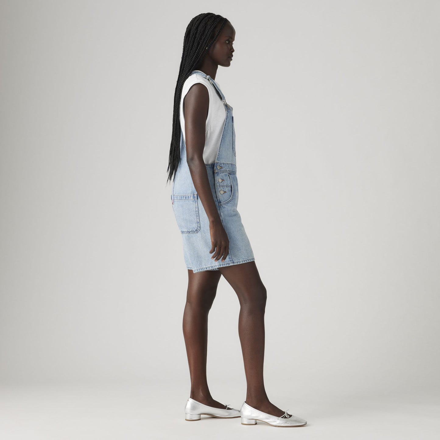 A woman in profile wears a white sleeveless top, Levi's Utility Shortall in relaxed-fit light blue denim with a mid-length cut, and white flats against a plain light background.