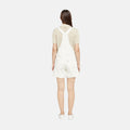 A woman faces away, wearing a cream lace short-sleeve top styled with Levi's Utility Shortall in white and matching white shoes, all set against a plain white background.