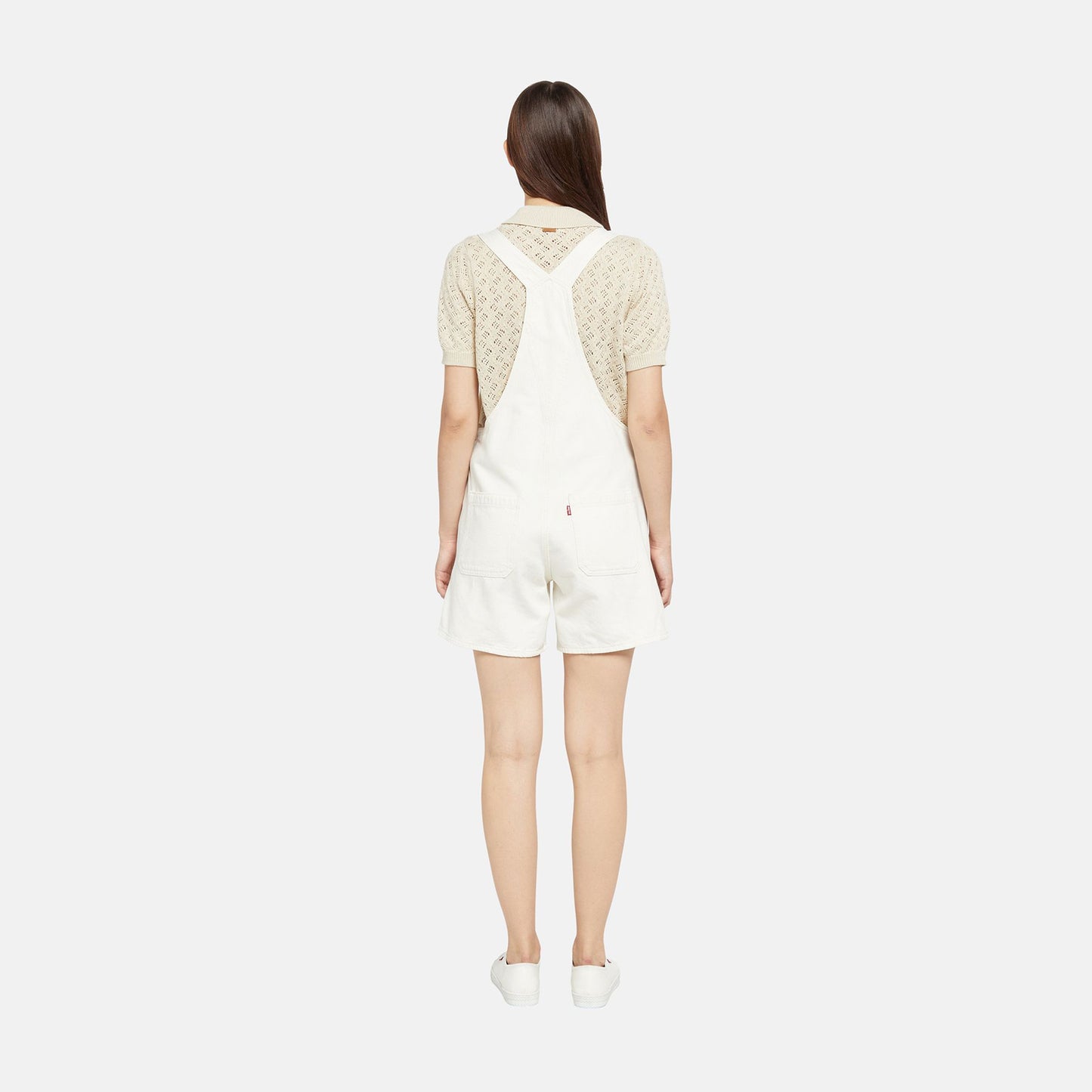 A woman faces away, wearing a cream lace short-sleeve top styled with Levi's Utility Shortall in white and matching white shoes, all set against a plain white background.