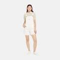 A woman stands against a plain background wearing Levis' Utility Shortall in white with a relaxed fit and mid-length cut, paired with a beige short-sleeved blouse and white sneakers.