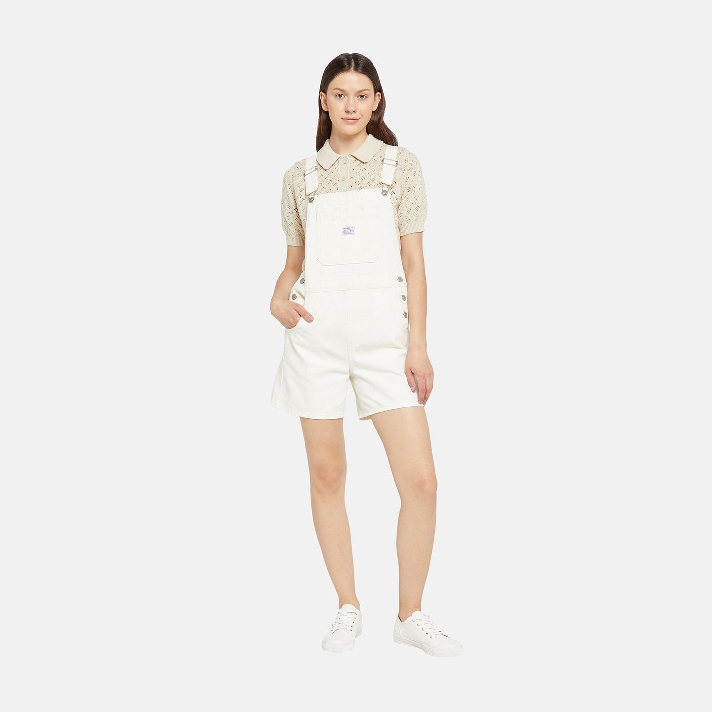 A woman stands against a plain background wearing Levis' Utility Shortall in white with a relaxed fit and mid-length cut, paired with a beige short-sleeved blouse and white sneakers.
