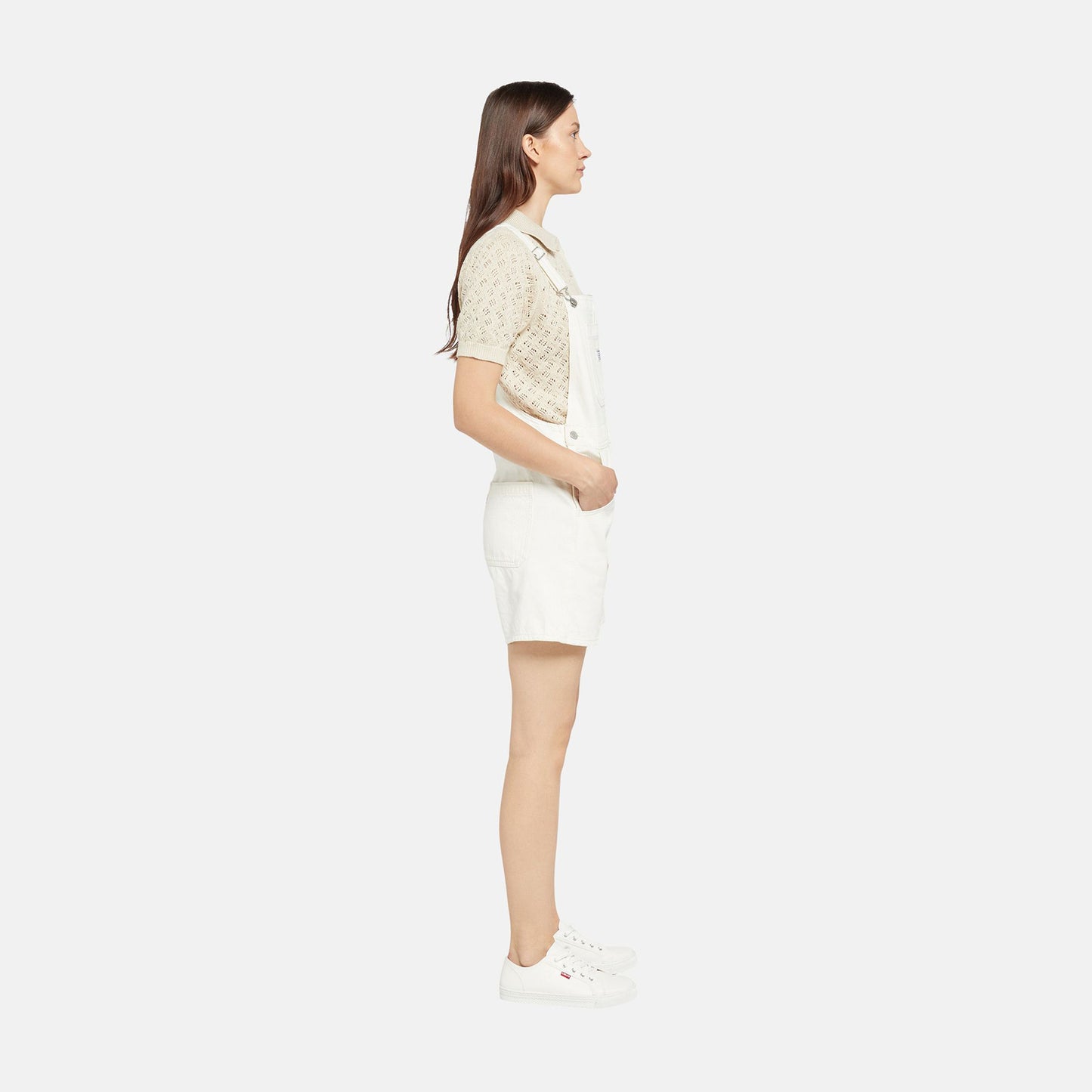 A woman stands in profile against a plain white background wearing the Levis Utility Shortall over a relaxed fit short-sleeve shirt and white sneakers.