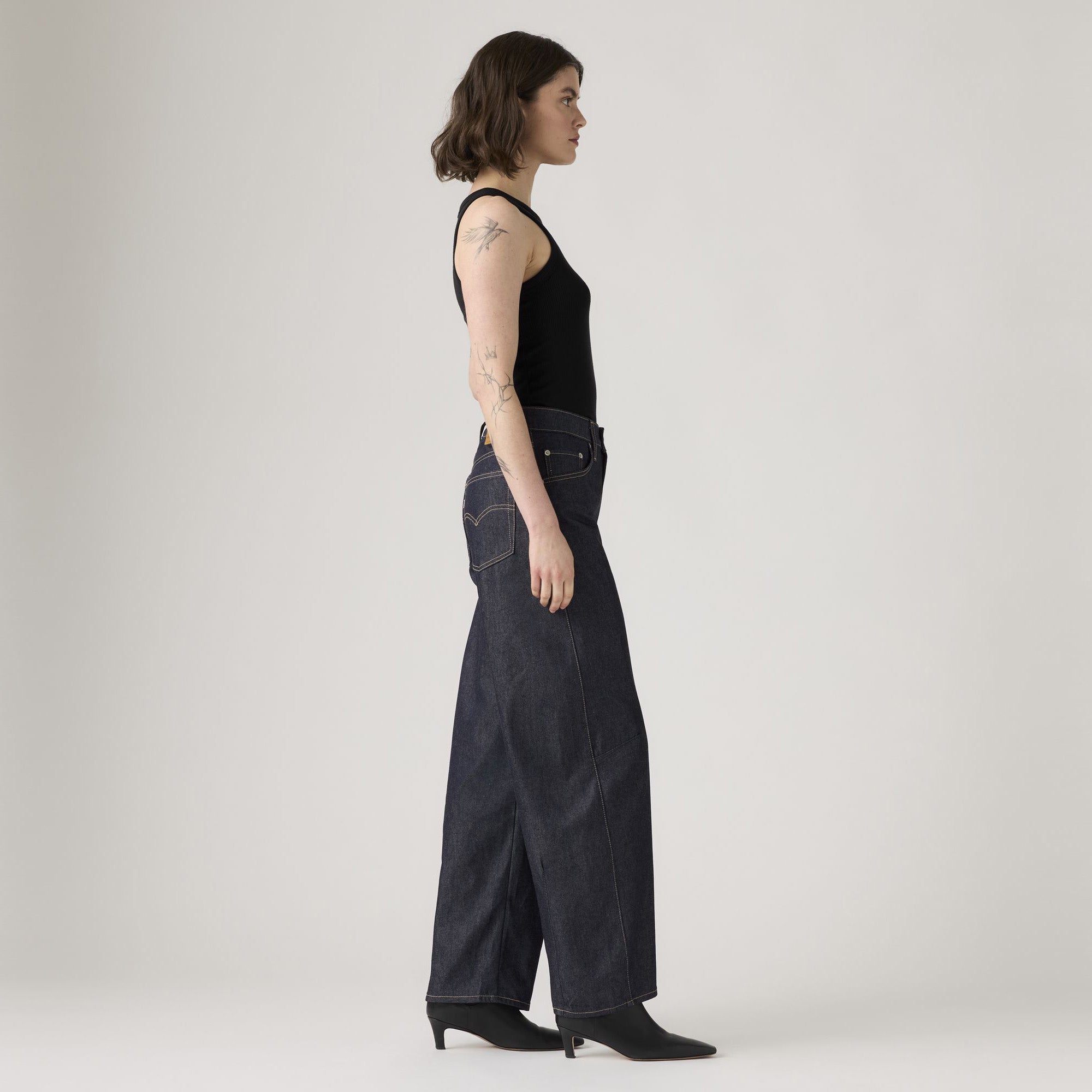 A woman stands in profile against a plain background, wearing a black tank top, Levis Baggy Dad Barrel jeans, and black ankle boots.