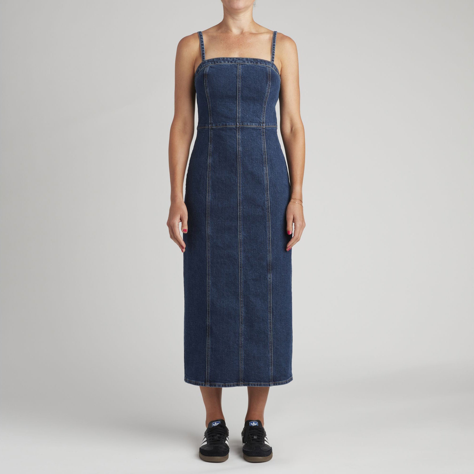 A person stands facing forward in the Levis Seamed Bustier Dress, an ankle-length denim piece with corset-inspired boning and thin straps, paired with black sneakers against a plain light background.