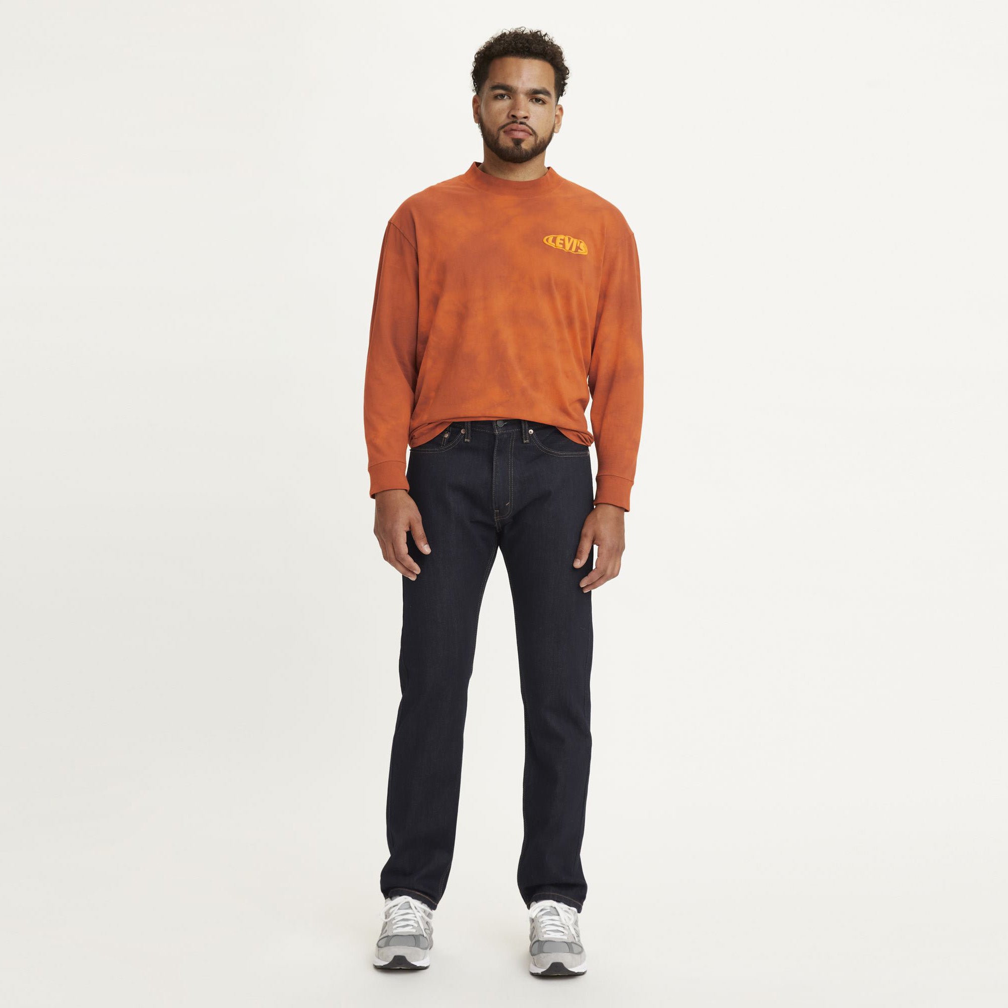 A man stands facing forward wearing a long-sleeve orange shirt, Levi's 505 Regular jeans, and gray sneakers against a plain white background.