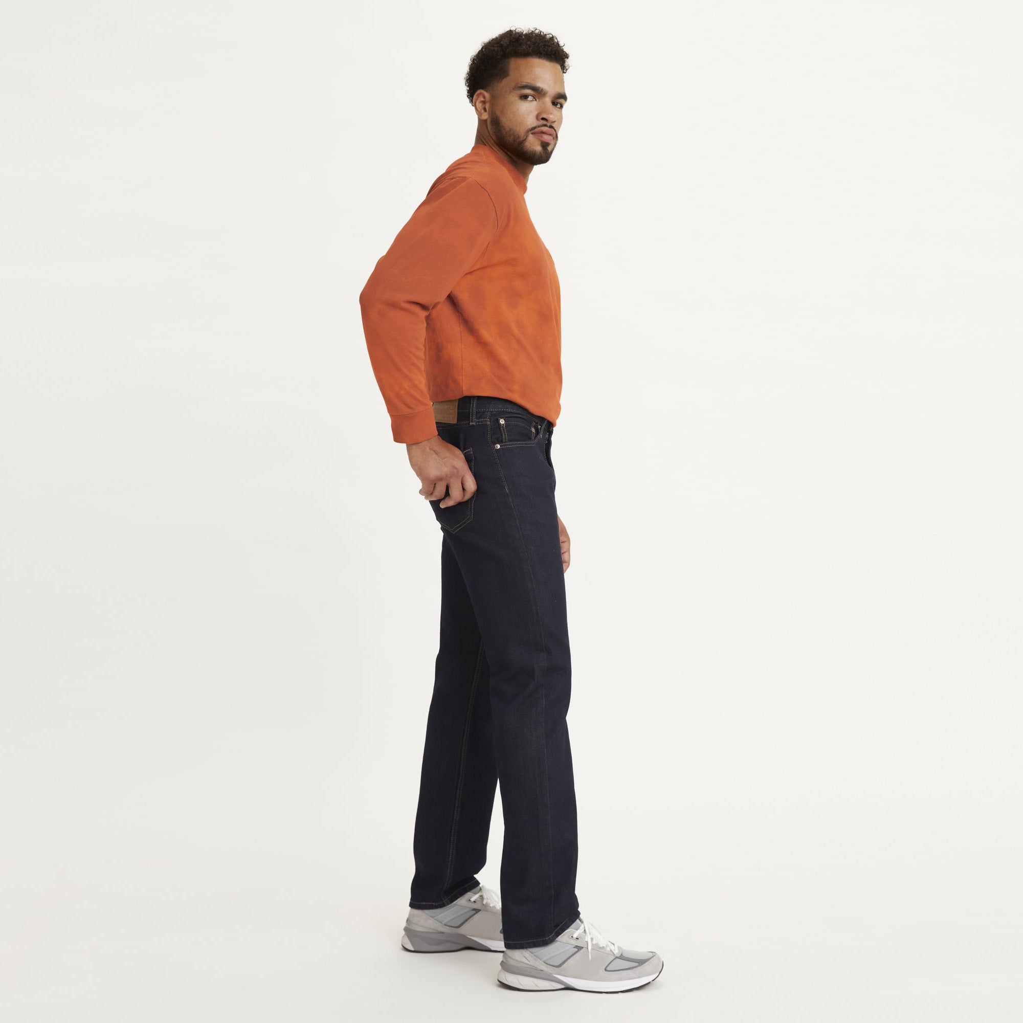 A man stands in profile wearing an orange long-sleeve shirt, Levi's 505 Regular stretch denim jeans, and gray sneakers against a plain white background.