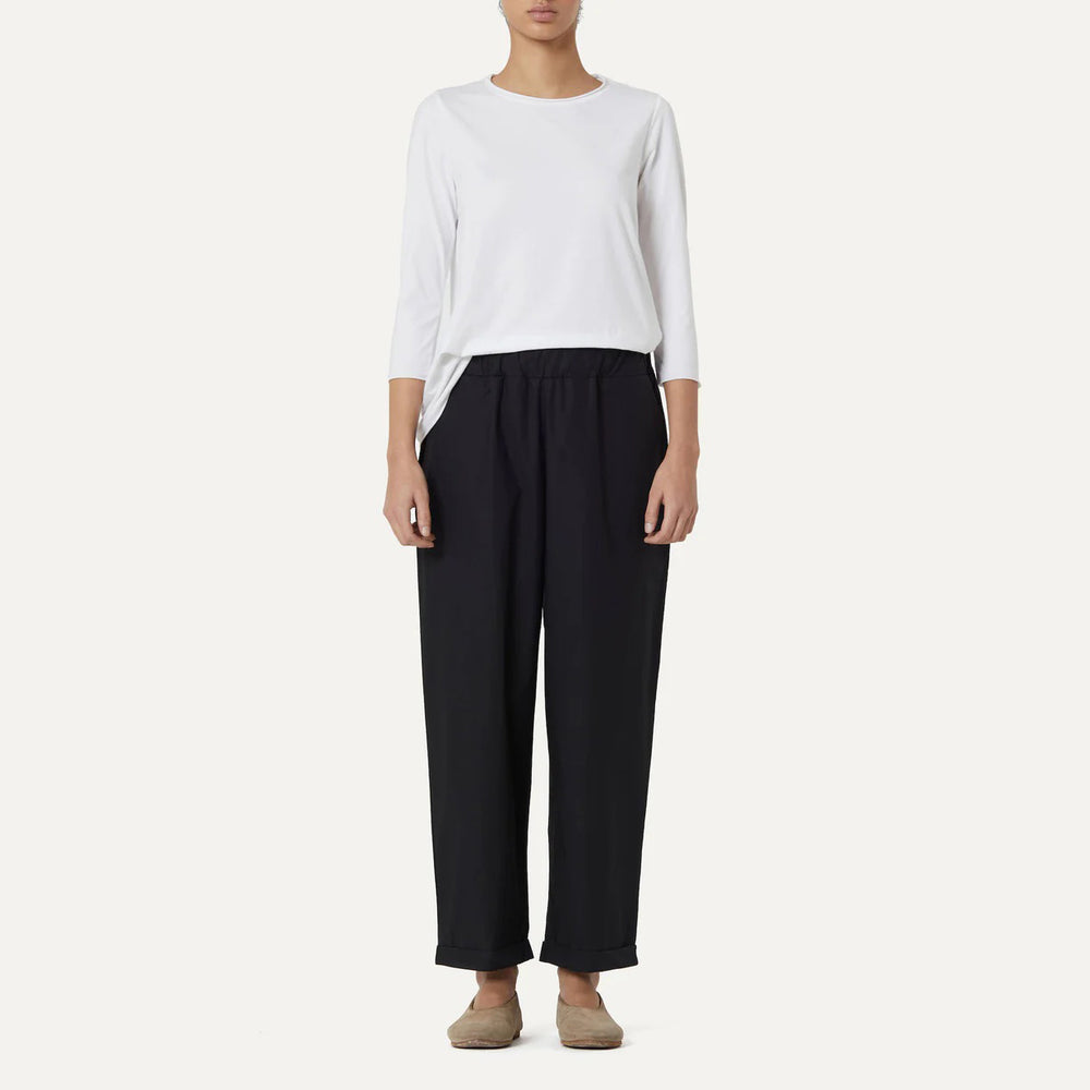 A person dressed in a white cotton poplin long-sleeve shirt, Labo Art's Panta Vela Pant, Clara in black with an elastic waistband, and beige flat shoes stands against a plain light background.
