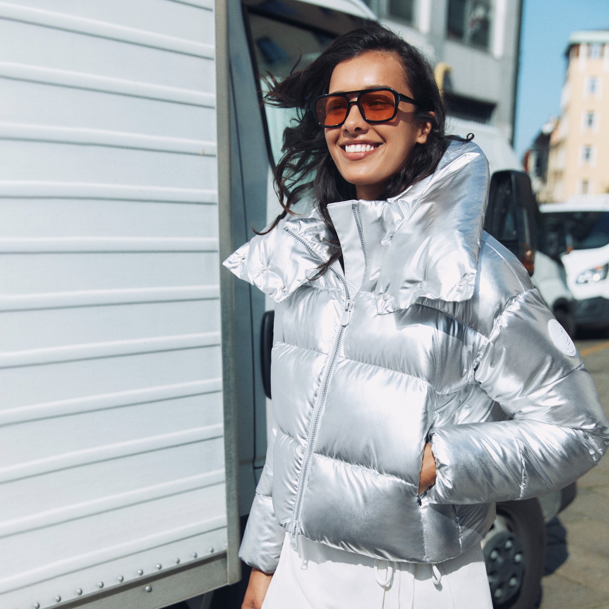 Woman wearing a silver puffer jacket and sunglasses standing next to a vehicle.