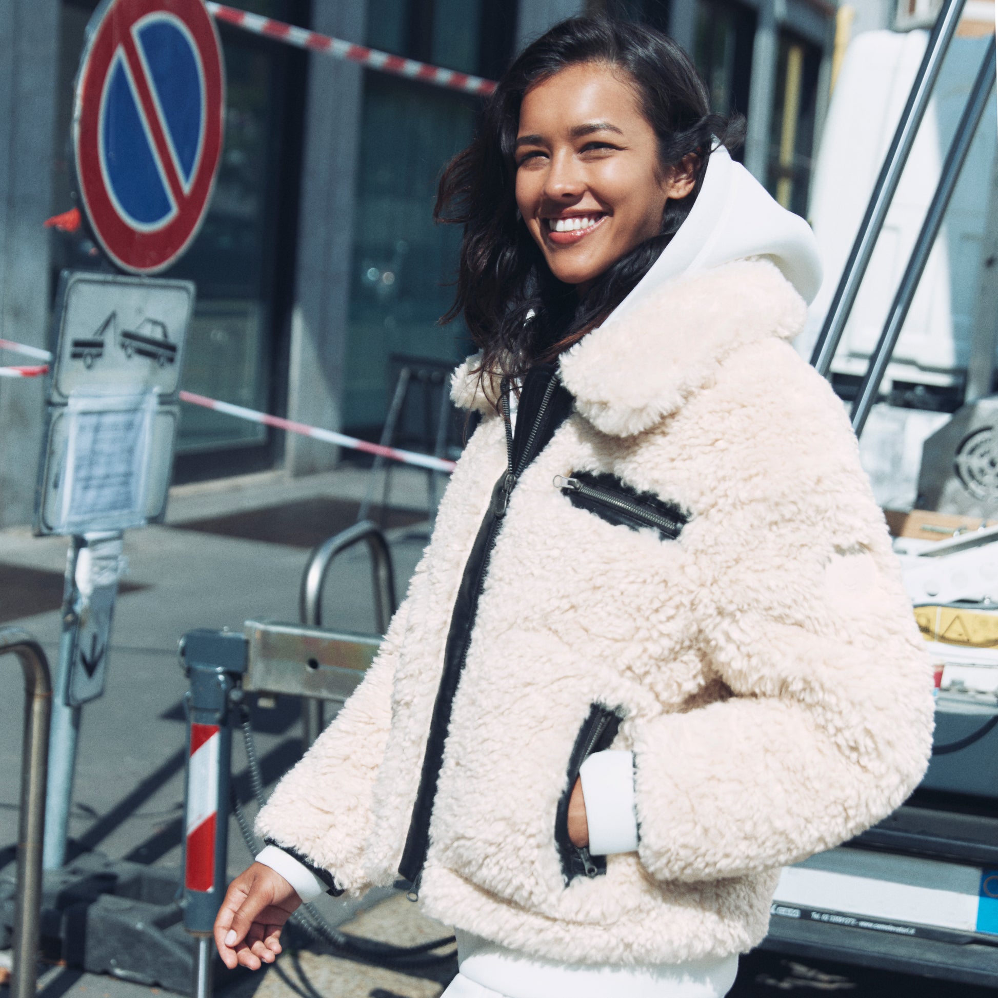 Woman wearing a beige sherpa jacket standing outdoors near a no parking sign.