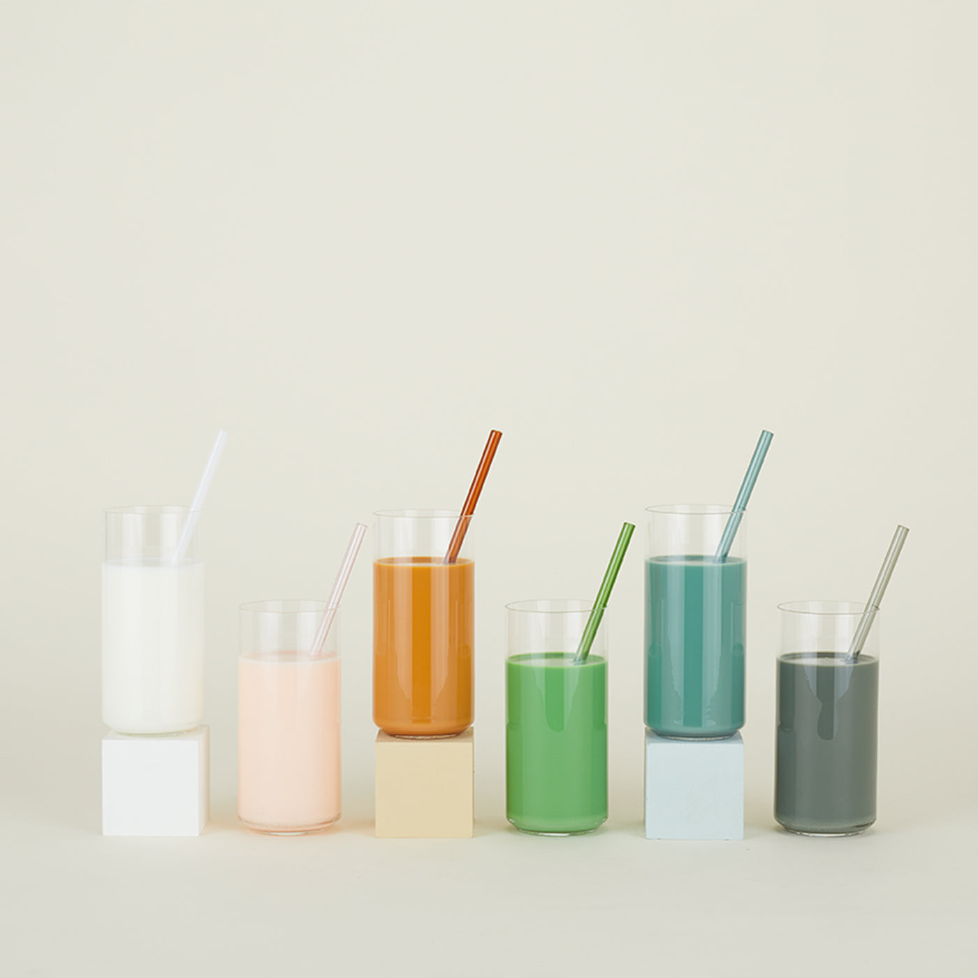 The Essential Glass Straws - Set of 6 by Hawkins New York includes six borosilicate glass tumblers with colored liquids and matching straws on small blocks. This eco-friendly arrangement adds a vibrant touch to any setting against a light background.