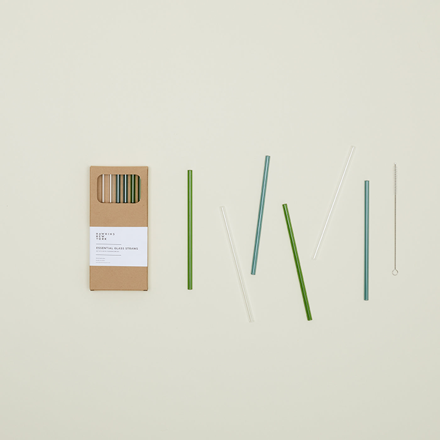 The Essential Glass Straws from Hawkins New York, including a cleaning brush and packaged in a cardboard box, are elegantly arranged on a light background.