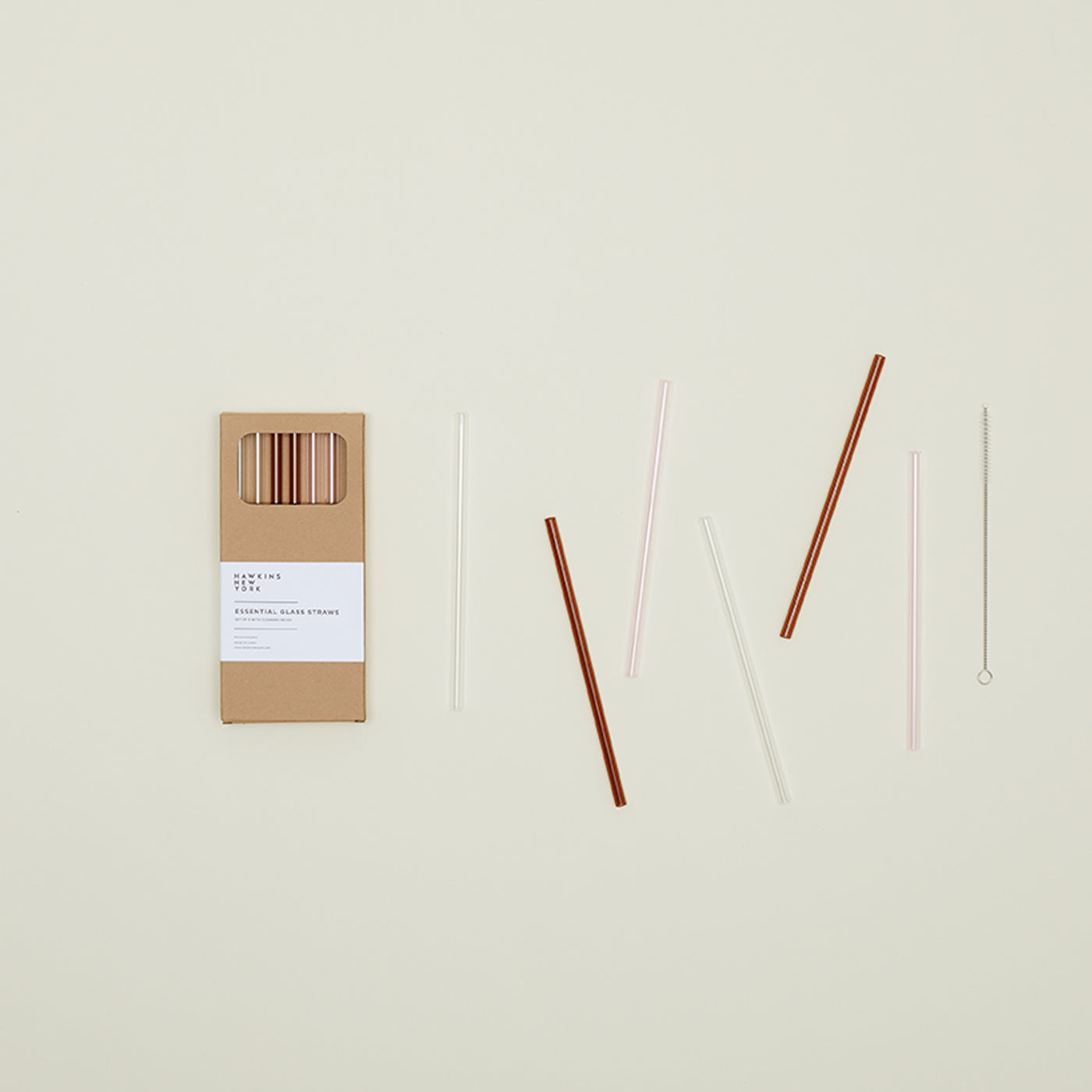 The Essential Glass Straws - Set of 6 by Hawkins New York, made from eco-friendly borosilicate glass and complete with a cleaning brush, are elegantly presented next to a brown cardboard box on a light background.