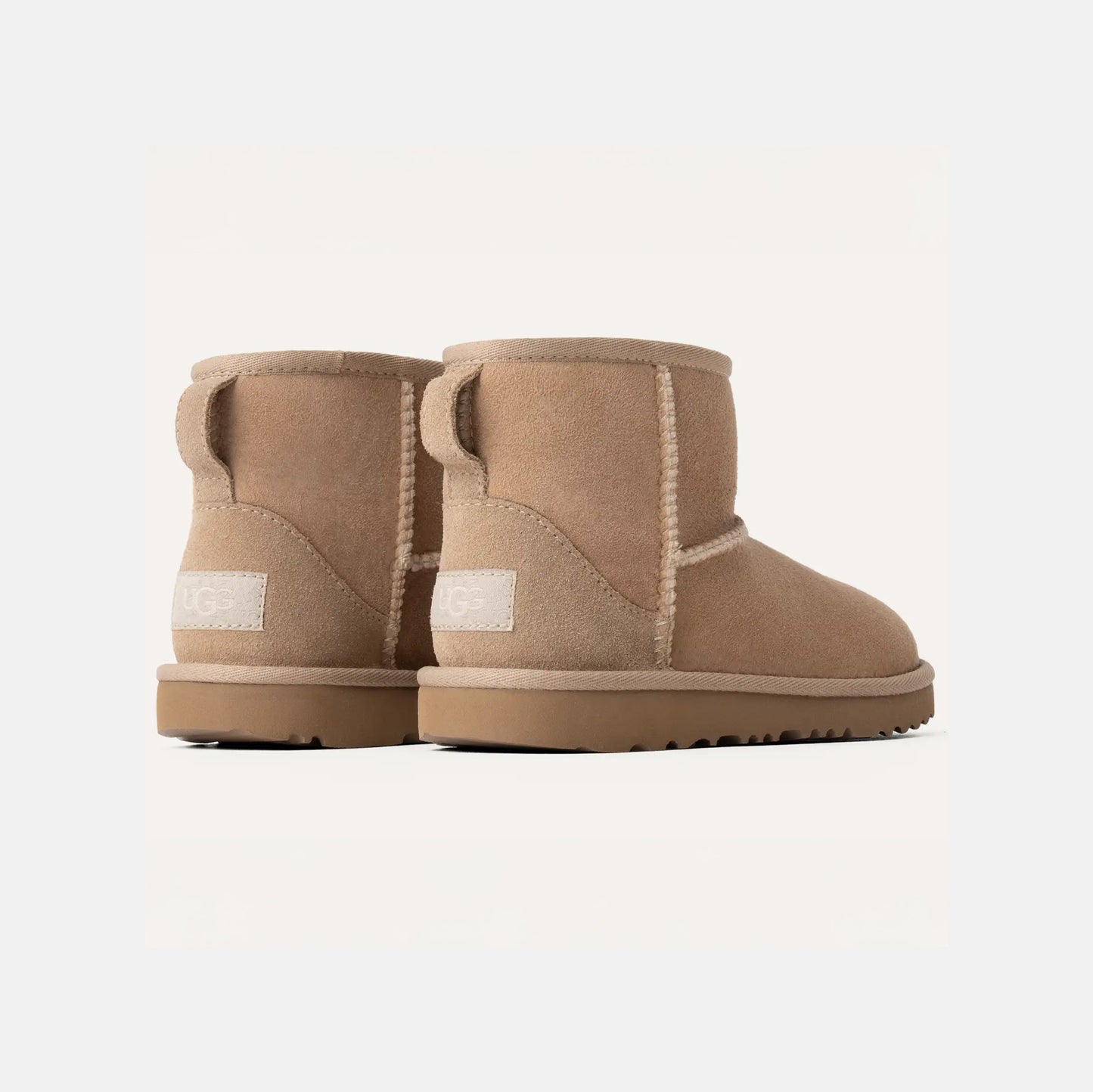 Two Ugg K Classic Mini II light brown suede boots with flat soles and visible stitching, featuring a cushioned outsole, are displayed back to back.