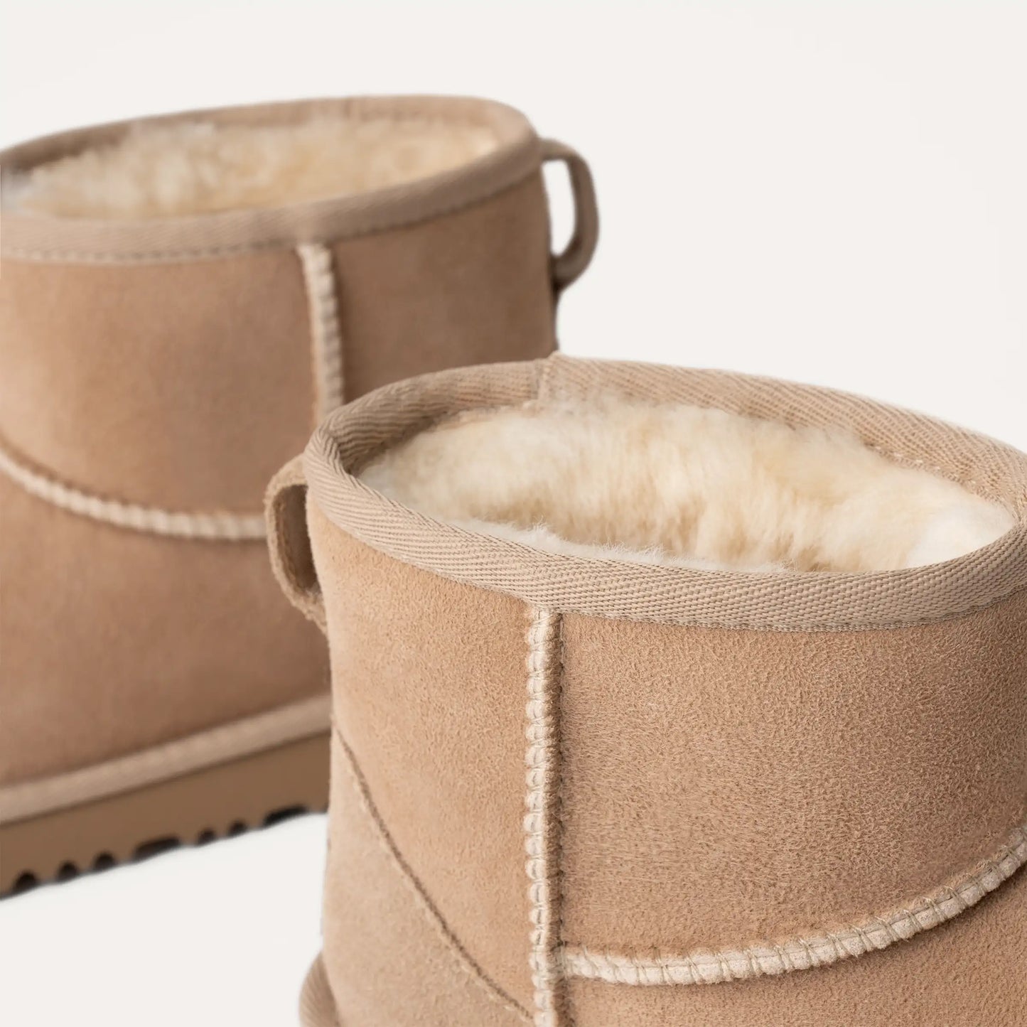 Two K Classic Mini II boots by Ugg, featuring tan suede sheepskin with fluffy interiors, cushioned outsoles, and looped back handles, are shown against a plain background.