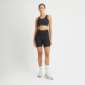 A person wears Rohnisch's Flattering High Waist Bike Shorts in black moisture-wicking fabric paired with white sneakers against a plain white background, hands resting by their sides.
