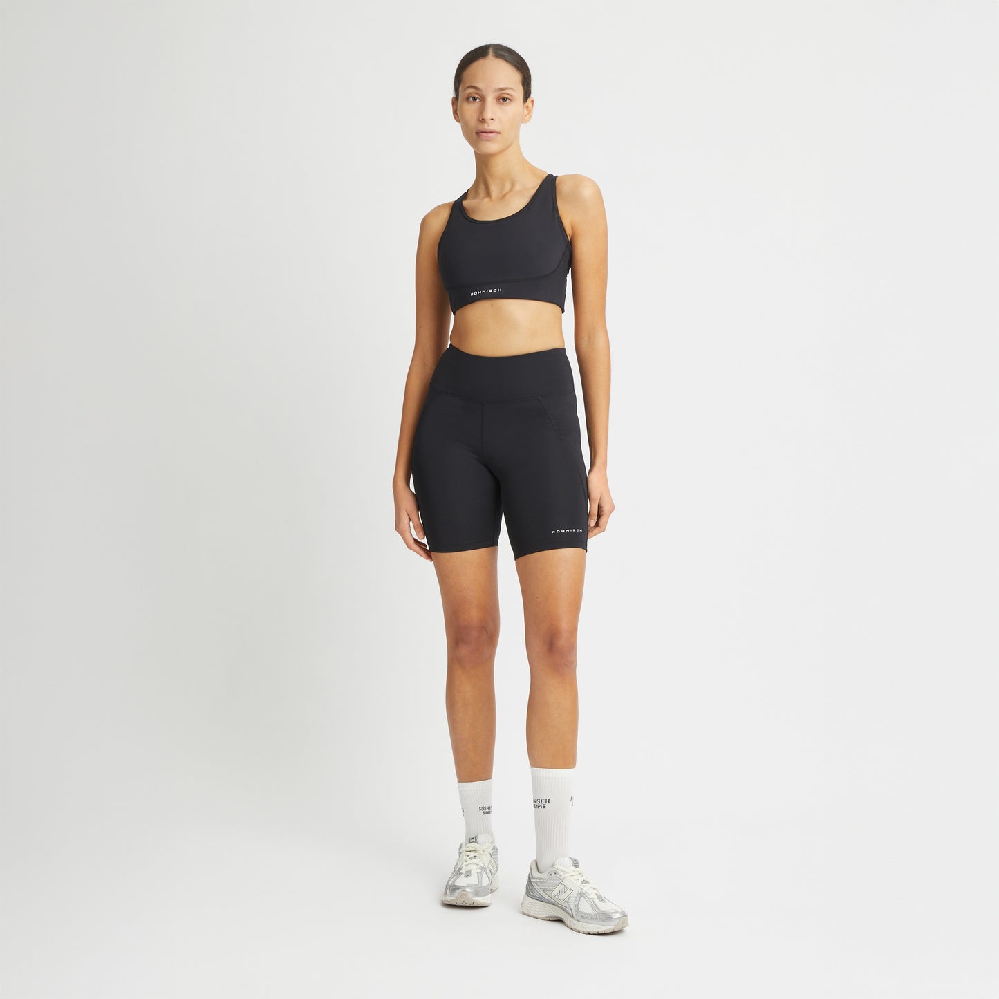 A person wears Rohnisch's Flattering High Waist Bike Shorts in black moisture-wicking fabric paired with white sneakers against a plain white background, hands resting by their sides.