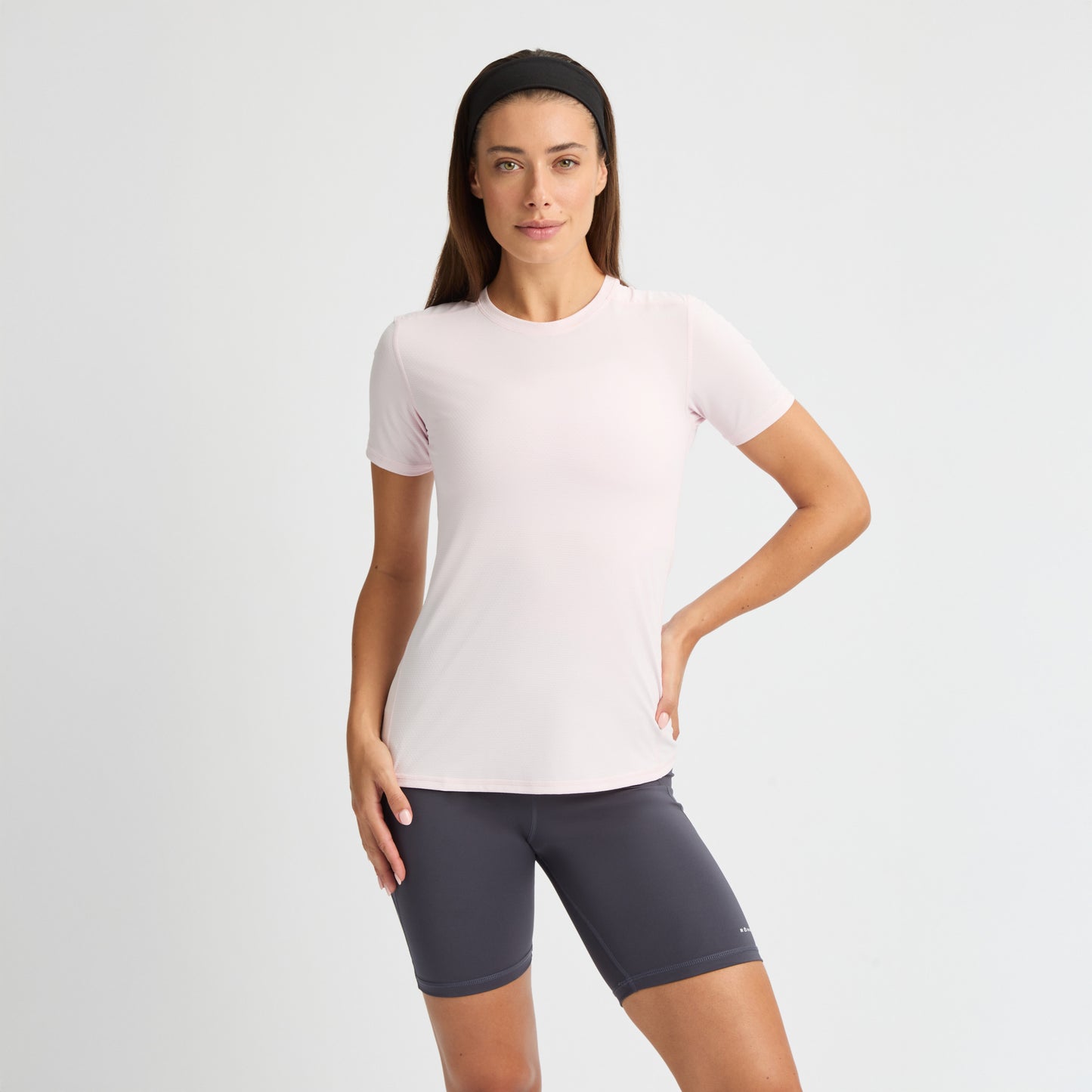 A person in a Jacquard Short Sleeve Tee by Rohnisch, made of breathable, recycled polyester, and dark shorts stands confidently with one hand on their hip against a plain background.