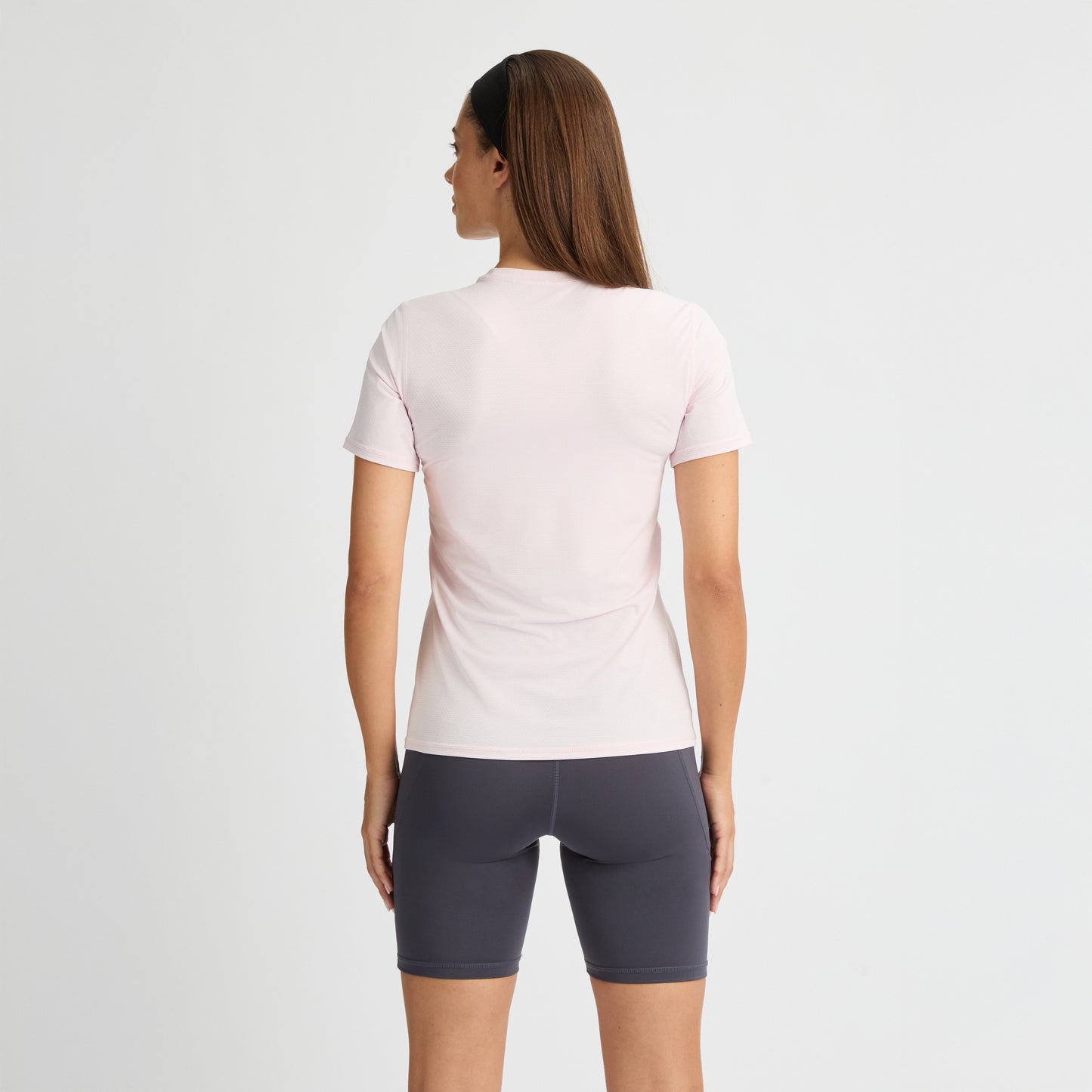 A woman with long hair, facing away, wears a Rohnisch Jacquard Short Sleeve Tee in pale pink made from breathable, recycled polyester paired with dark gray shorts against a white background.
