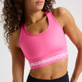A person with curly hair and a neutral expression wears the Rohnisch Emblem Racerback Sportsbrah in pink, featuring white text.