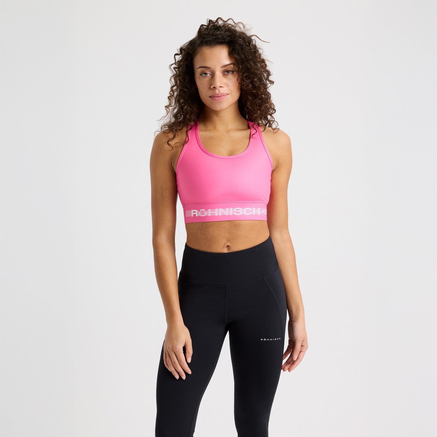 A person in a pink Emblem Racerback Sportsbrah by Rohnisch and black leggings, crafted for high support and moisture-wicking performance, stands against a plain white background.