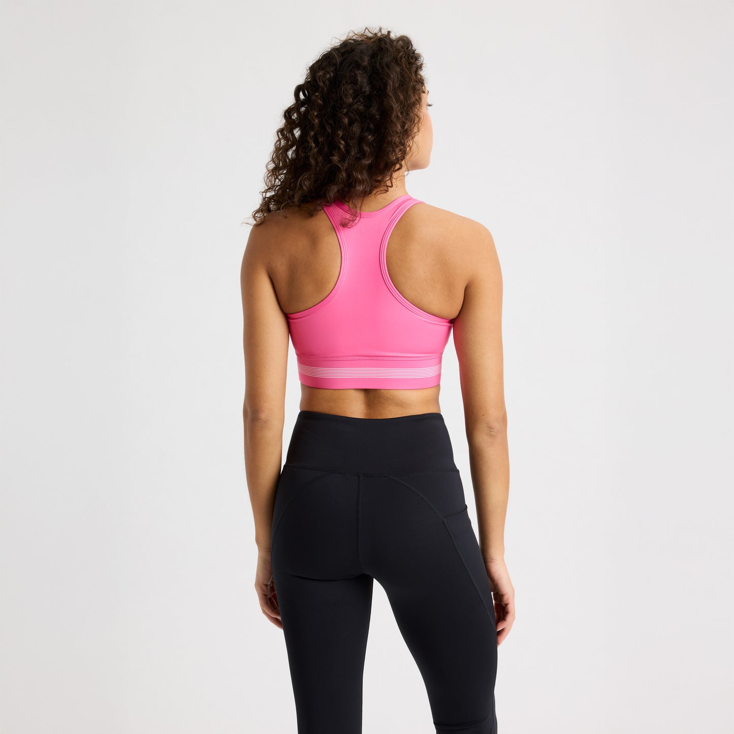 A person with curly hair stands with their back to the camera, wearing a pink Emblem Racerback Sportsbrah by Rohnisch paired with black leggings.
