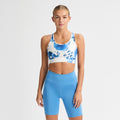 A person wearing a Rohnisch Emblem Racerback Sportsbrah in moisture-wicking blue and white, paired with blue shorts, stands against a plain white background.