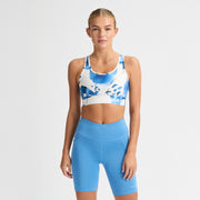A person wearing a Rohnisch Emblem Racerback Sportsbrah in moisture-wicking blue and white, paired with blue shorts, stands against a plain white background.