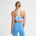 A woman with a ponytail models Rohnisch's Emblem Racerback Sportsbrah, featuring high support and moisture-wicking technology, in blue and white. She stands against a plain backdrop with matching blue leggings, shown from the back.