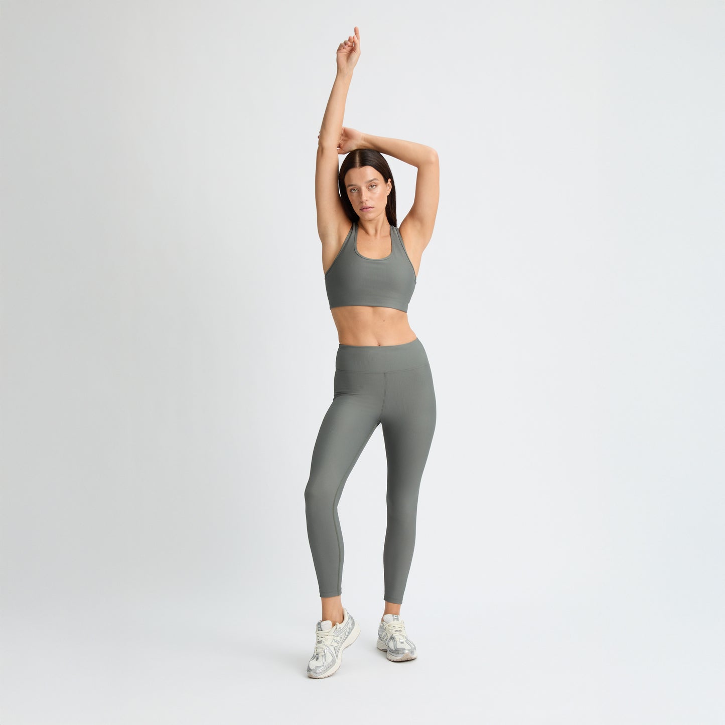 Wearing Rohnisch's Ribbed Regular High Waist Tights made from recycled polyester, a person in a gray sports bra and leggings stretches with arms raised, complemented by white sneakers against a plain white background.