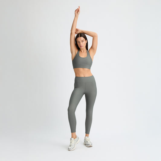 Wearing Rohnisch's Ribbed Regular High Waist Tights made from recycled polyester, a person in a gray sports bra and leggings stretches with arms raised, complemented by white sneakers against a plain white background.