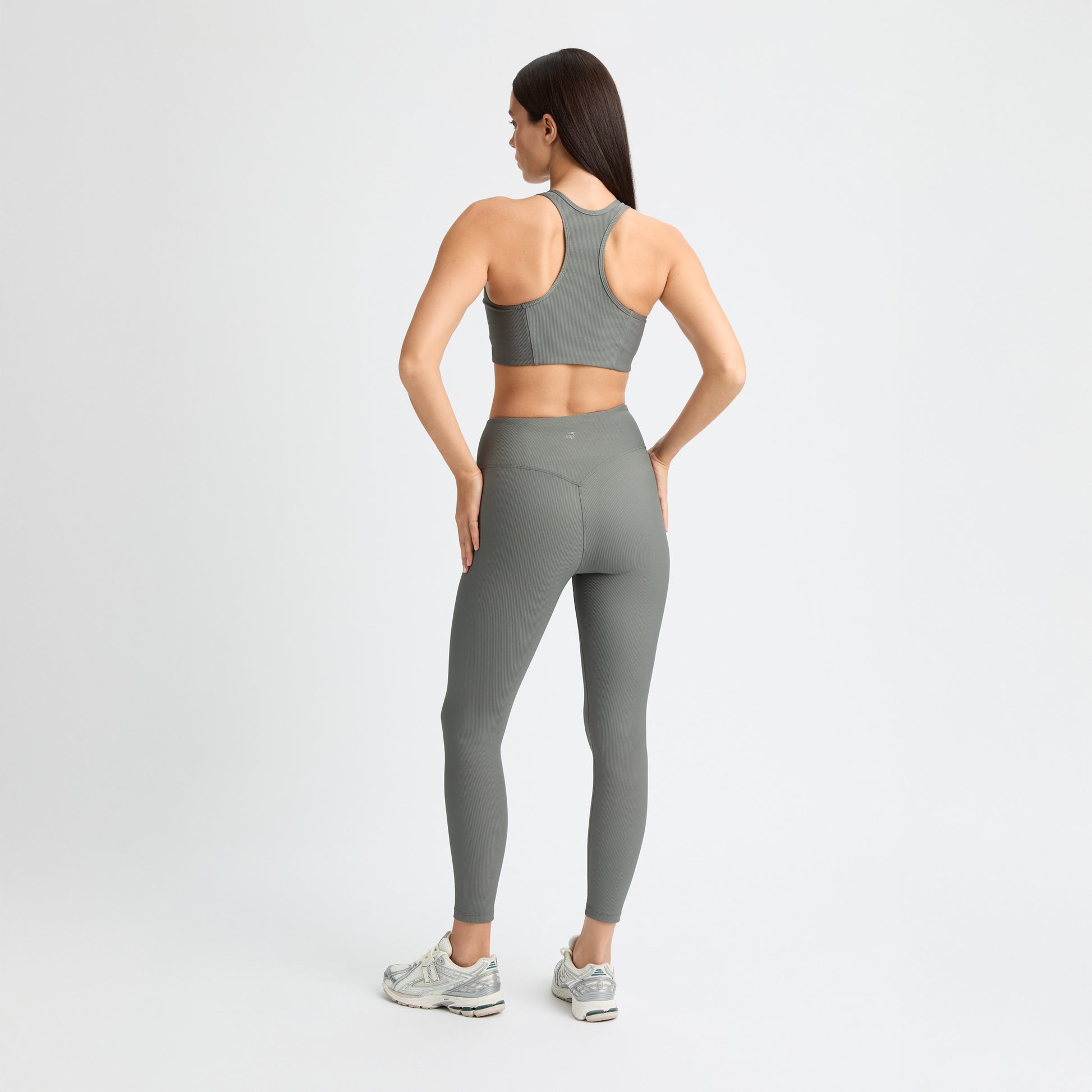 A woman in a matching gray workout outfit and sneakers, featuring Rohnisch Ribbed Regular High Waist Tights made from recycled polyester, stands with her hands on her hips against a plain white background.